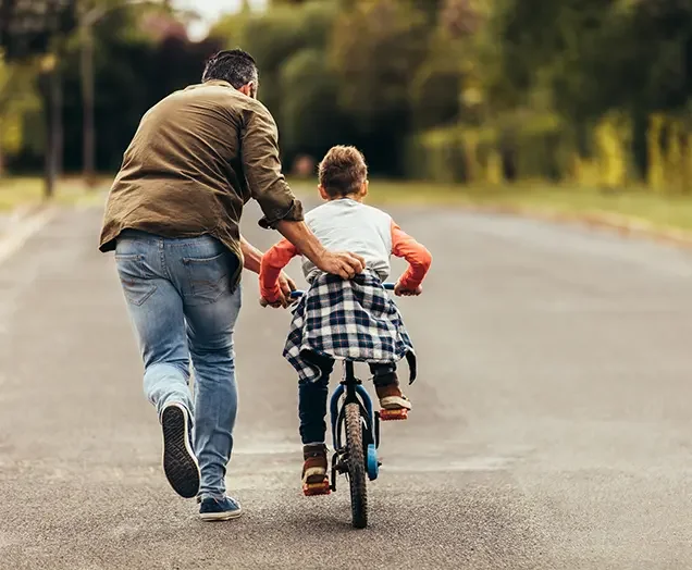 A man helping a young boy ride a bicycle on a paved road in a park with trees in the background.