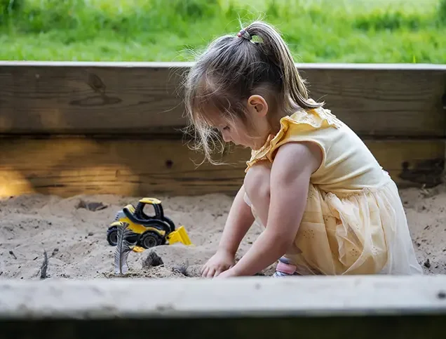 A young girl playing in a sandbox outside with toy construction vehicles.