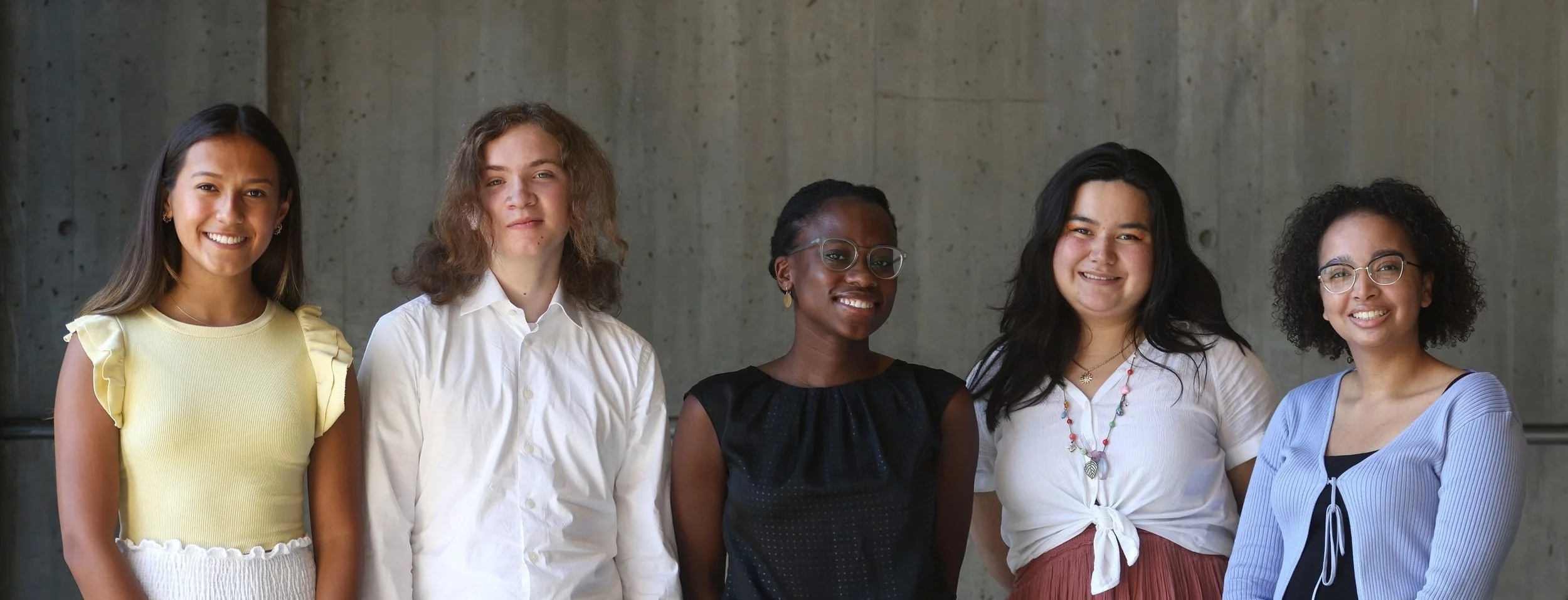 five young people stand in a line, smiling in front of a concrete wall in Boston City Hall.