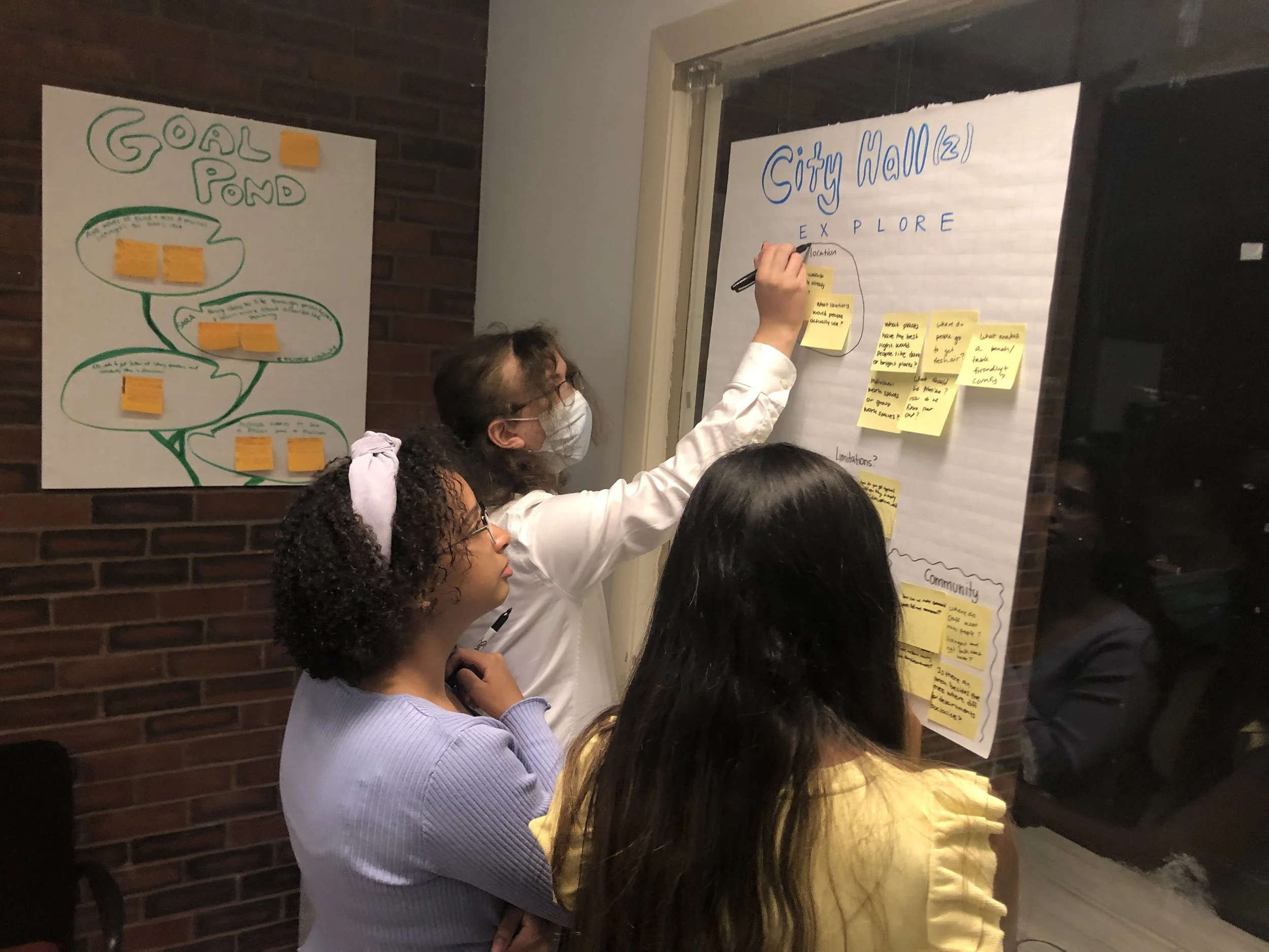 three young people stand in a small room looking at a poster on the wall. The poster says "City Hall, Explore". One person circles three post-it notes on the poster.
