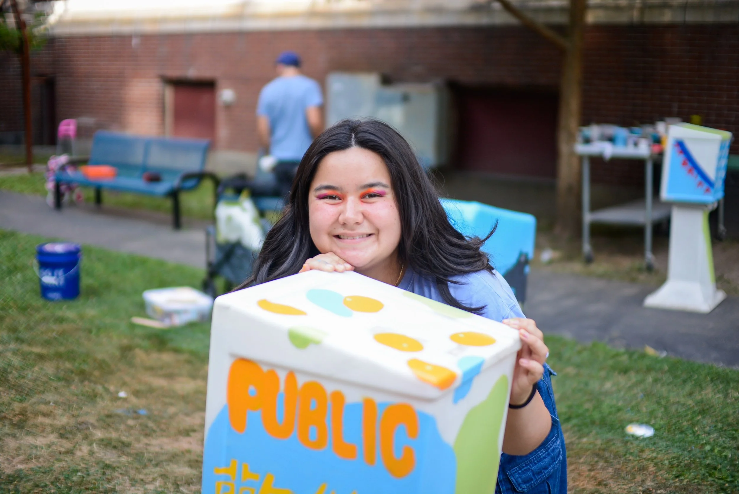 ava smiles holding her painted newspaper box. the box says PUBLIC in orange, round letters.
