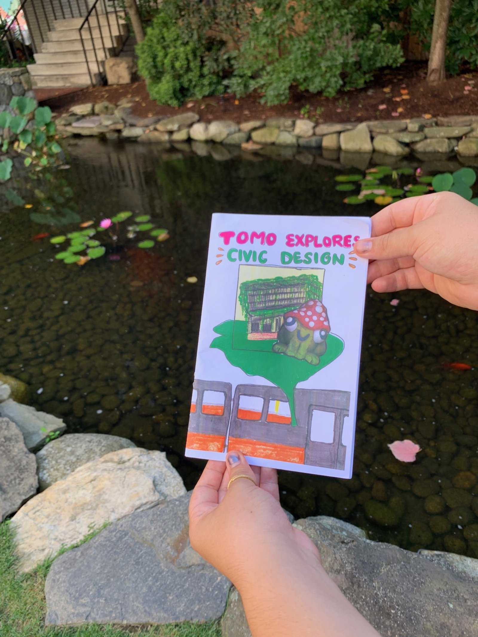 two hands hold a small paper zine above a pond. the cover says, "tomo explores civic design" and shows a frog character next to Boston City Hall.