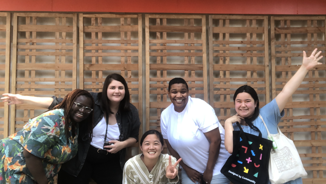 five young adults stand outside in front of a wooden wall. They are all smiling and standing in a line.