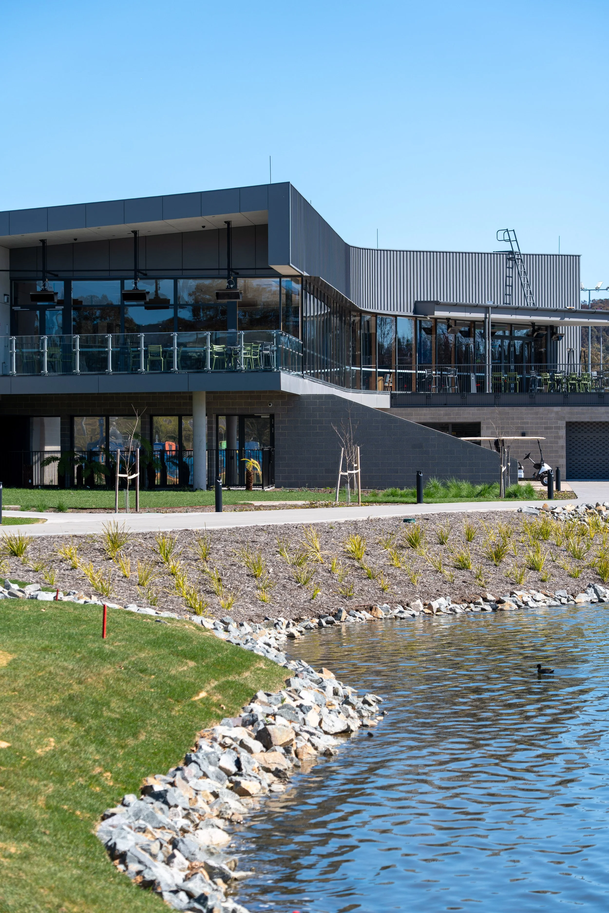 Modern building with large glass windows and a balcony overlooking a water body and landscaped lawn.