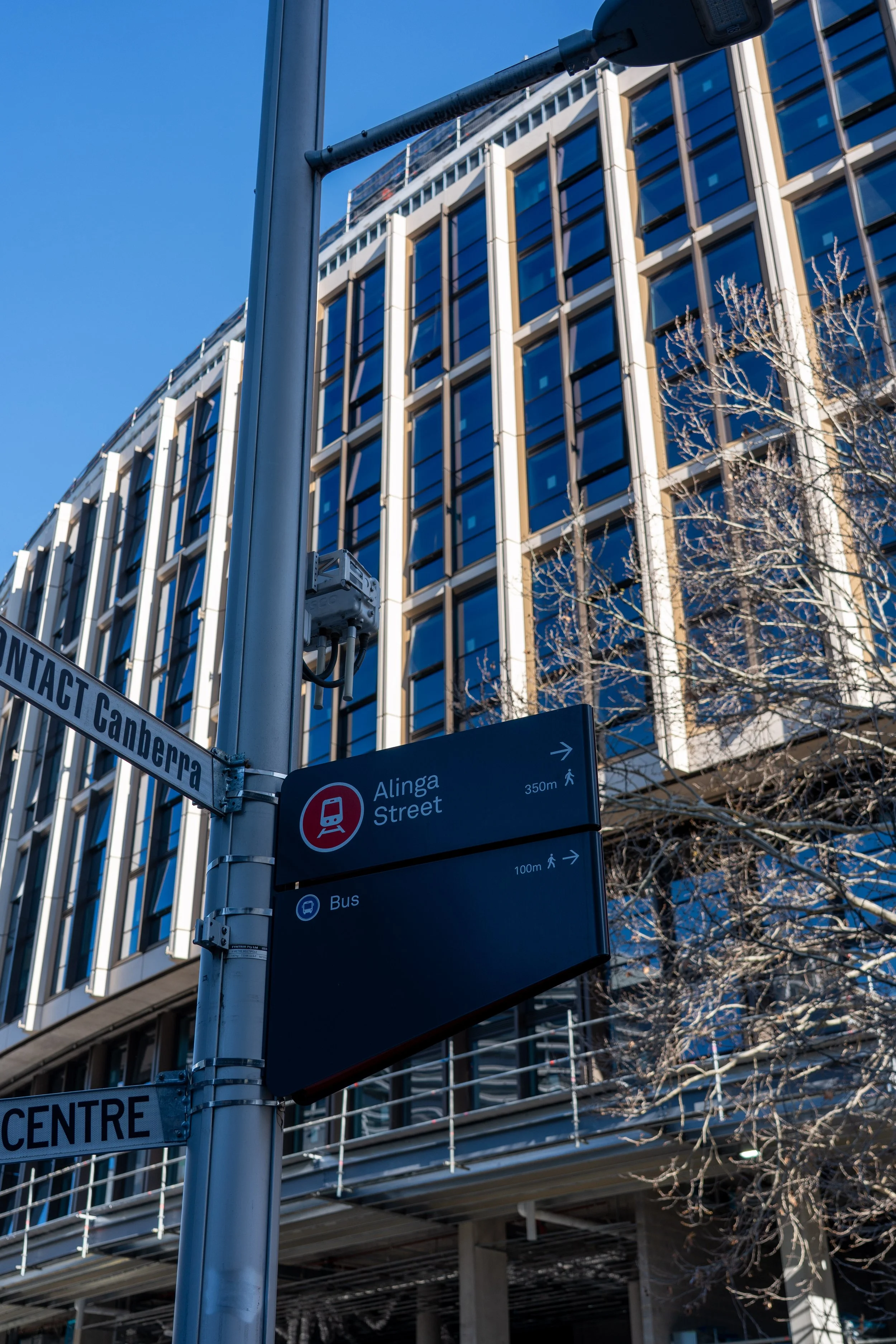 Street signs in front of a modern glass building with blue windows, showing directions for Alinga Street, Gengeira, and bus routes, with leafless trees and blue sky.