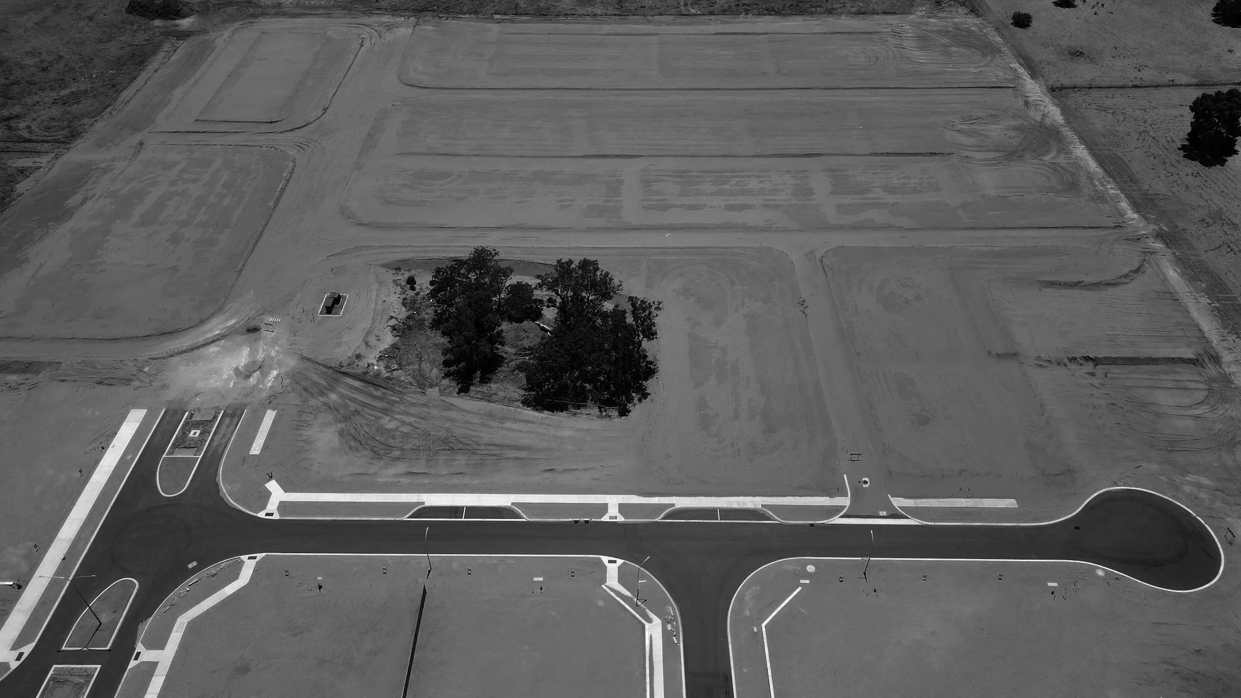 A black and white aerial view of an under-construction area with roads, open land, and a small cluster of trees.