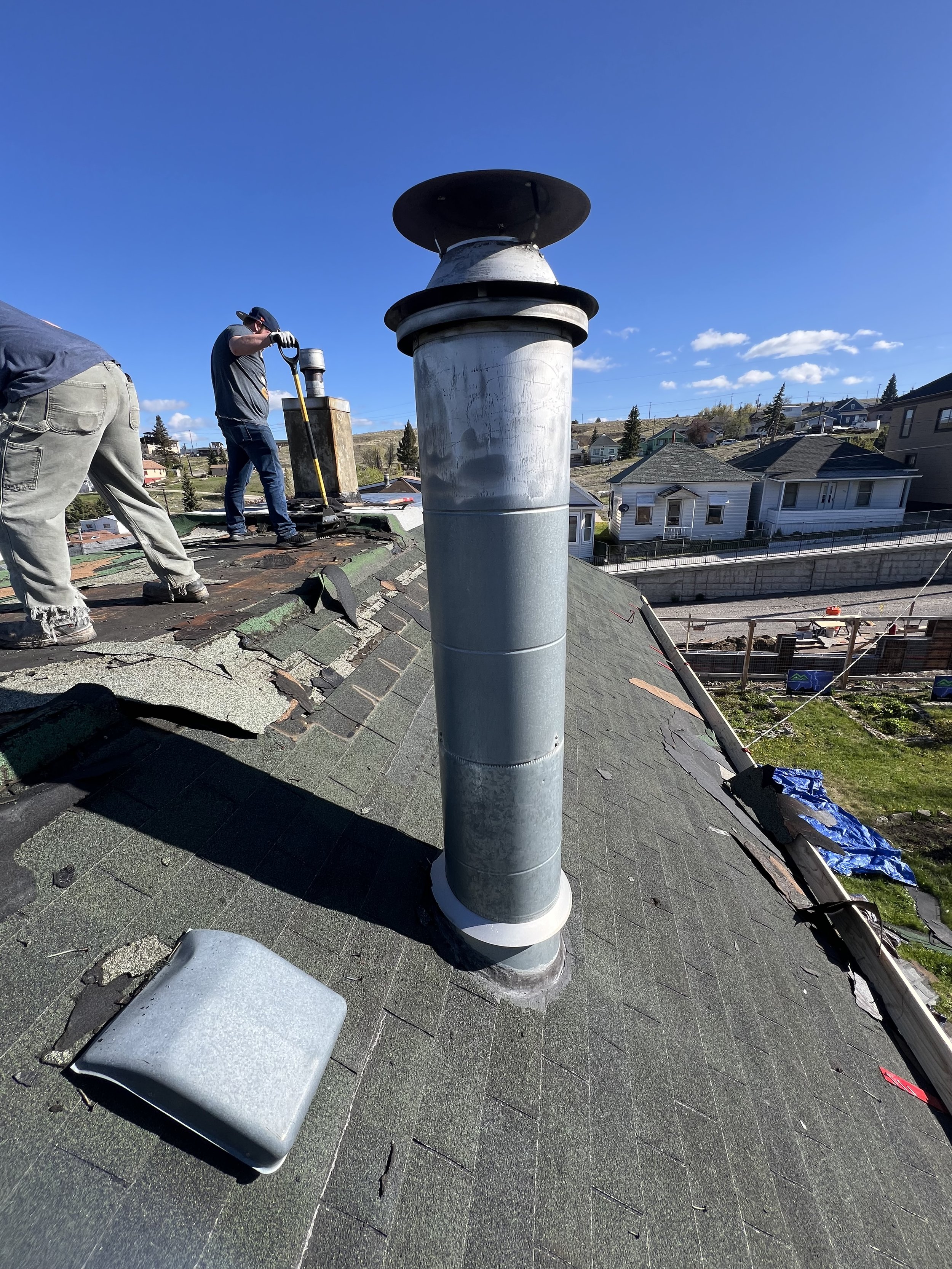 Roof with ridge vent, chimney pipe, and two workers repairing or installing the roofing shingles under a clear blue sky.