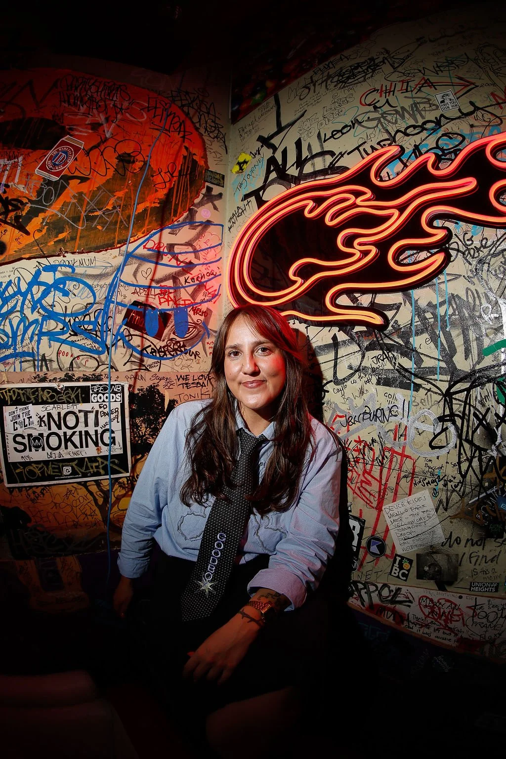A woman sitting in front of a graffiti-covered wall with a neon flame sign above her and a "No Smoking" sign on her left.