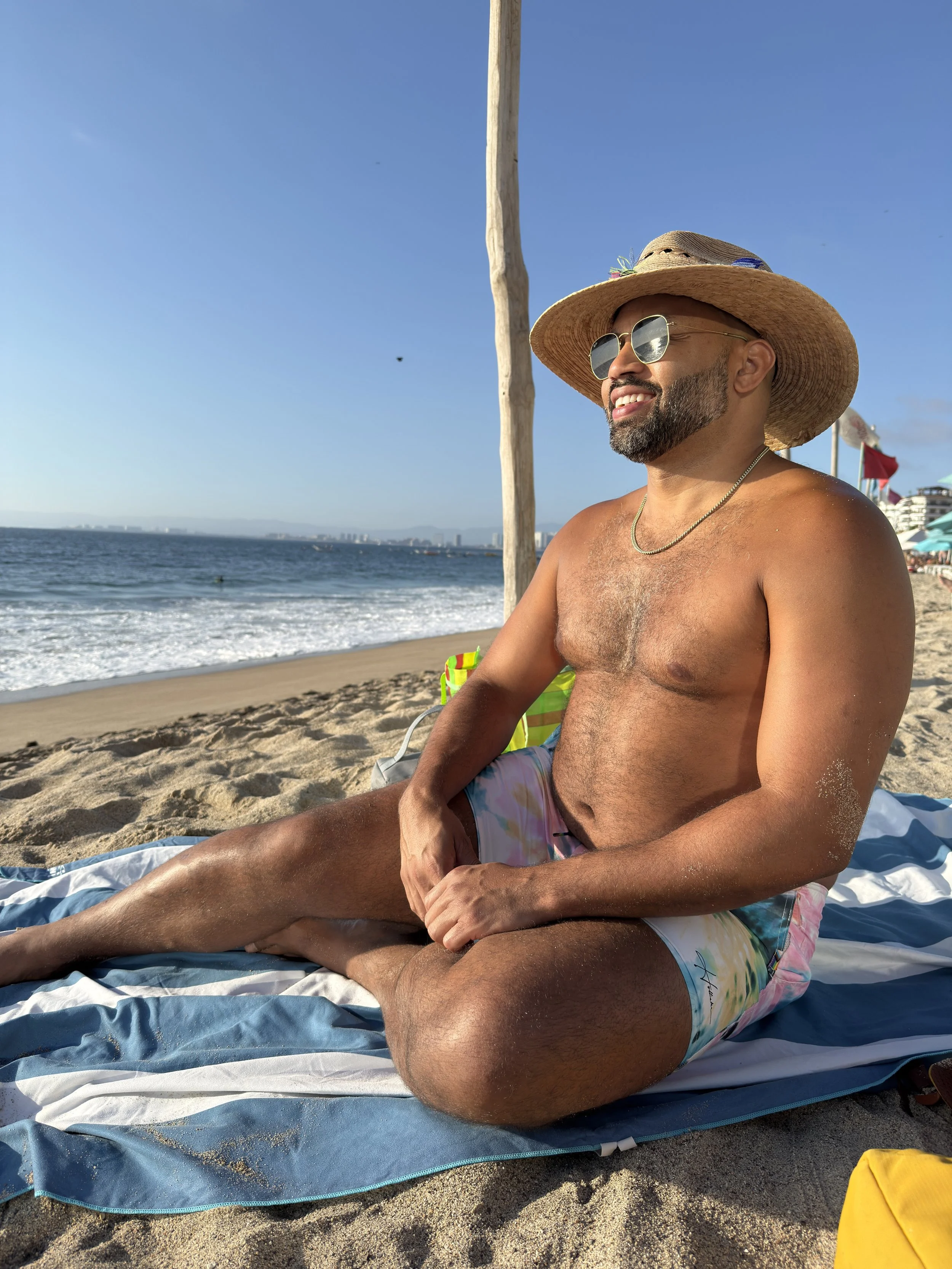 A man sitting on a beach in the sun, wearing a wide-brimmed hat, sunglasses, a gold necklace, and colorful swim trunks, with the ocean and a clear blue sky in the background.