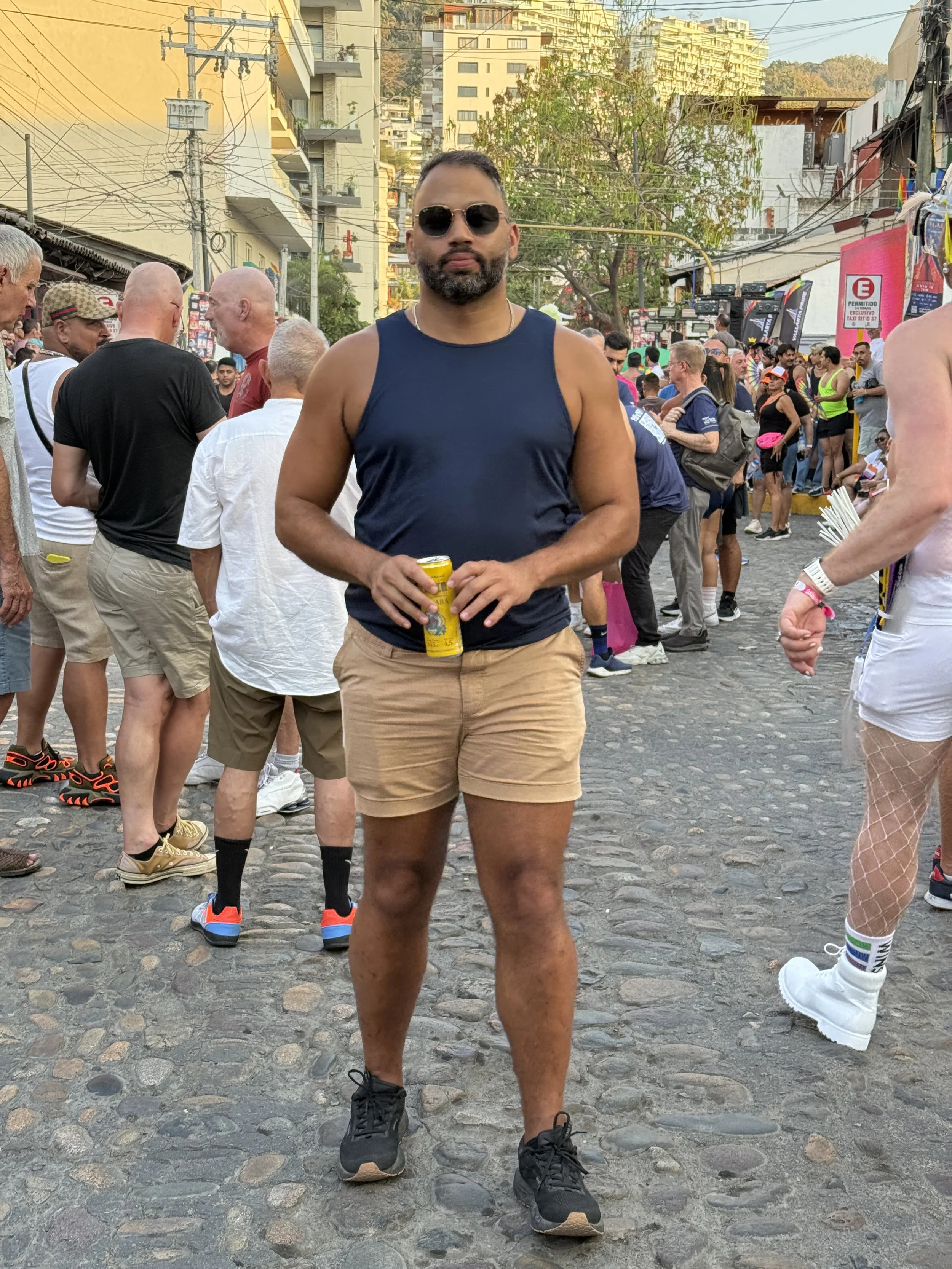 A man in a navy tank top, tan shorts, and black sneakers holding a yellow canned beverage, standing on a cobblestone street during a crowd event, wearing sunglasses, with a background of people and city buildings.