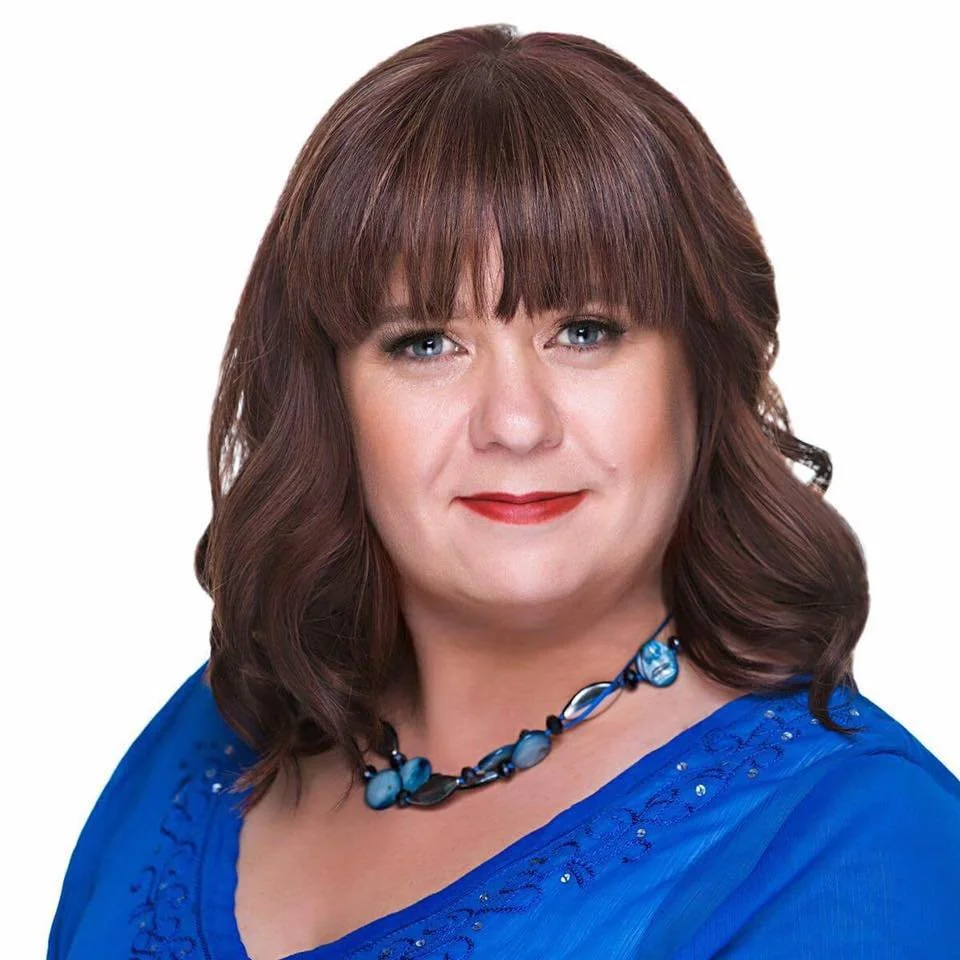 A woman with shoulder-length brown hair and bangs, wearing a blue top and a matching blue necklace, smiling against a white background.