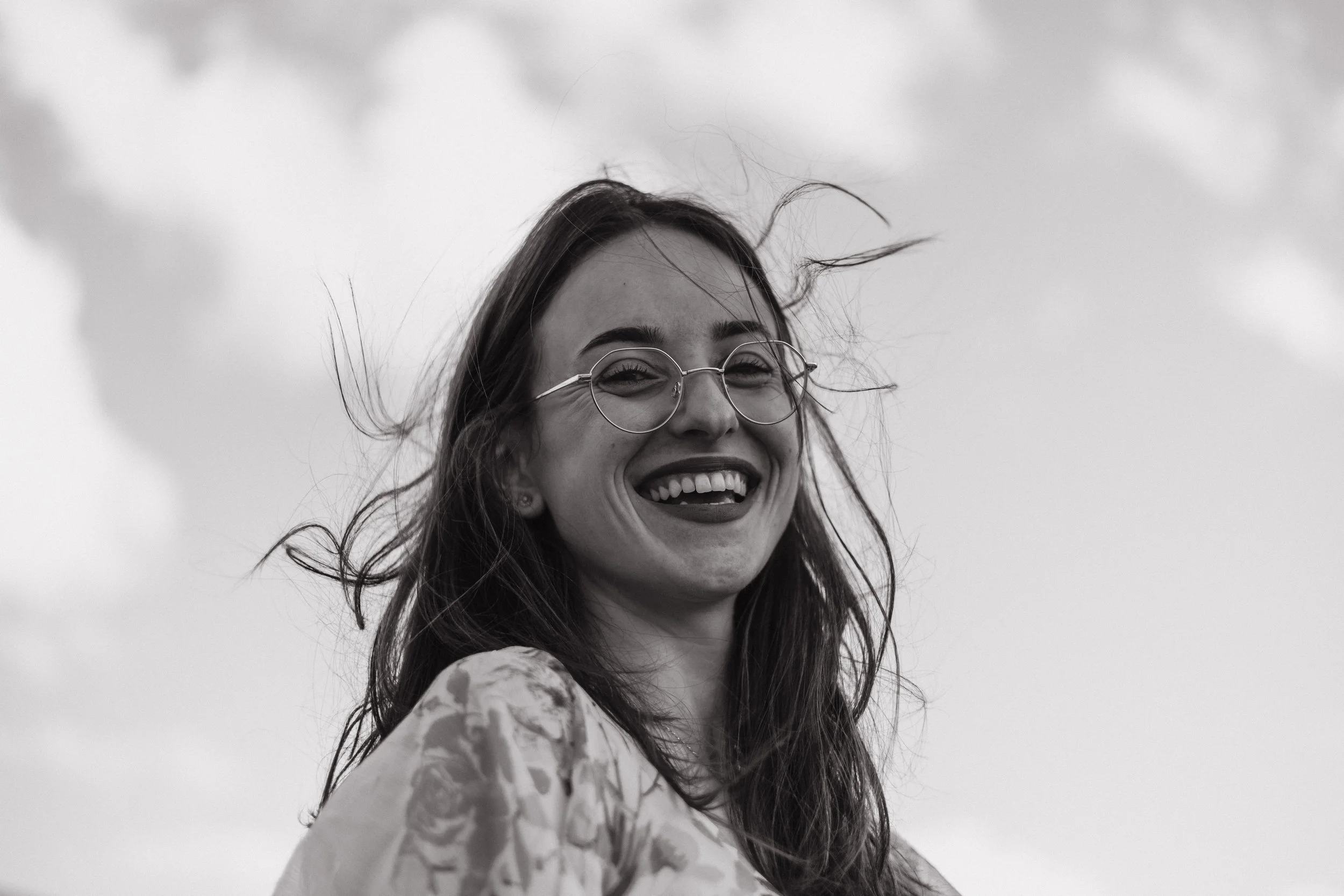 Black and white photo of a woman smiling outdoors with wind-blown hair, wearing glasses and a patterned top.