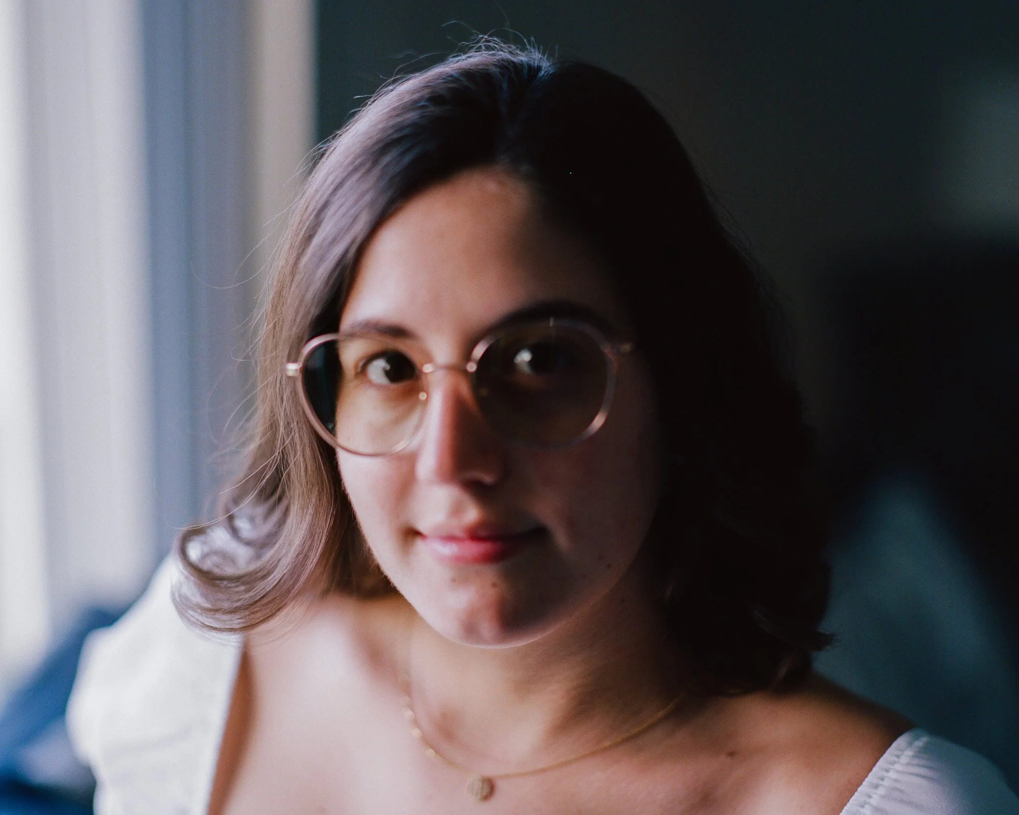 A young woman with shoulder-length dark hair wearing round glasses and a white top. She is looking directly at the camera with a slight smile, near a window with natural light.