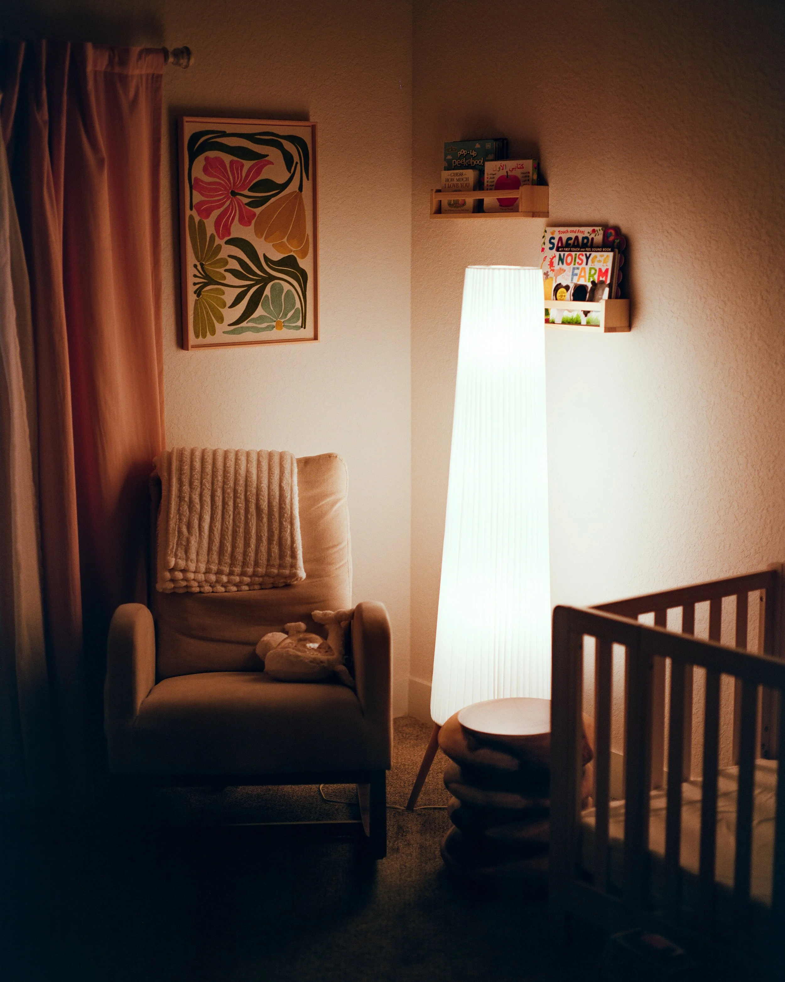 A cozy corner of a room with a beige armchair with a plush blanket and stuffed animal, a tall white floor lamp, a crib with a mobile, and wall shelves with children's books.