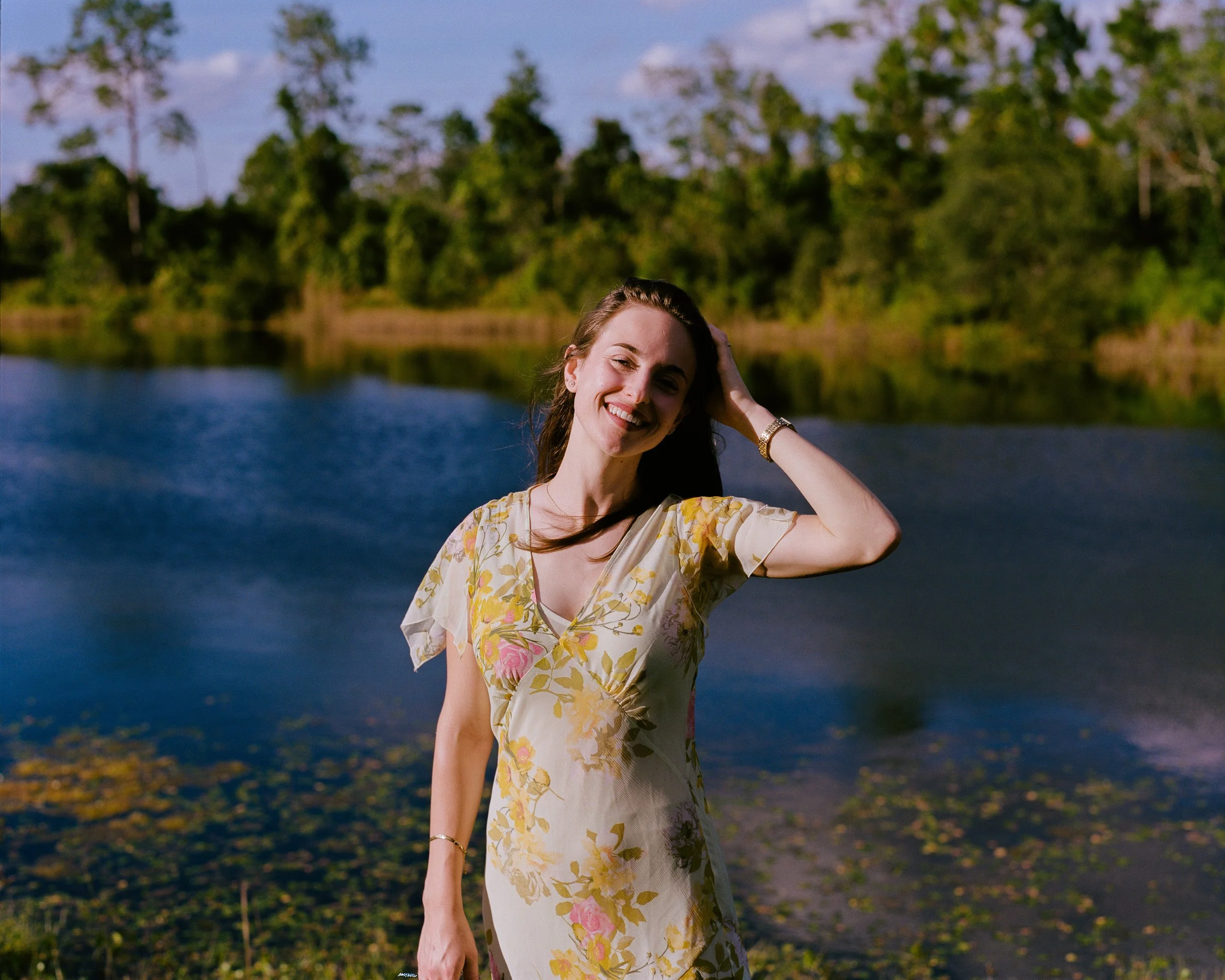 A woman smiling and posing by a river on a sunny day with lush green trees in the background.