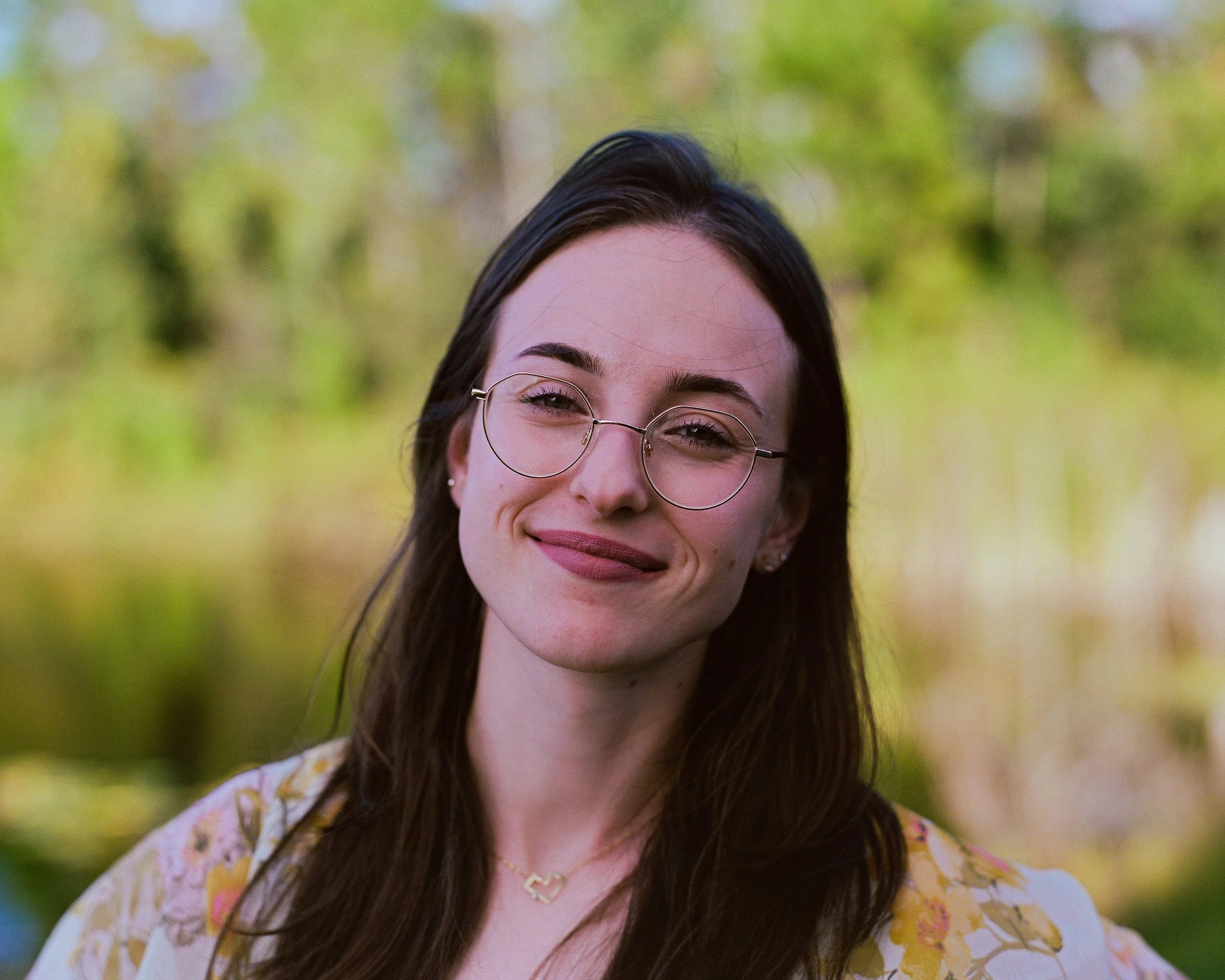 A young woman with long dark hair, round glasses, and a smile, standing outdoors with a blurred green and yellow background.