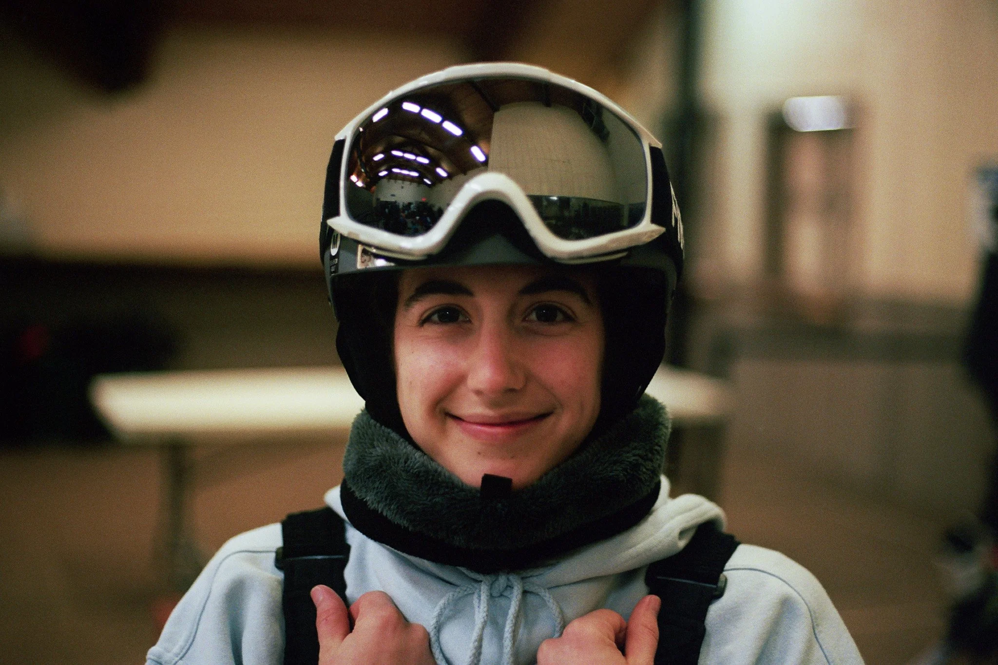 Young person wearing a ski helmet and goggles, smiling at the camera, indoors.