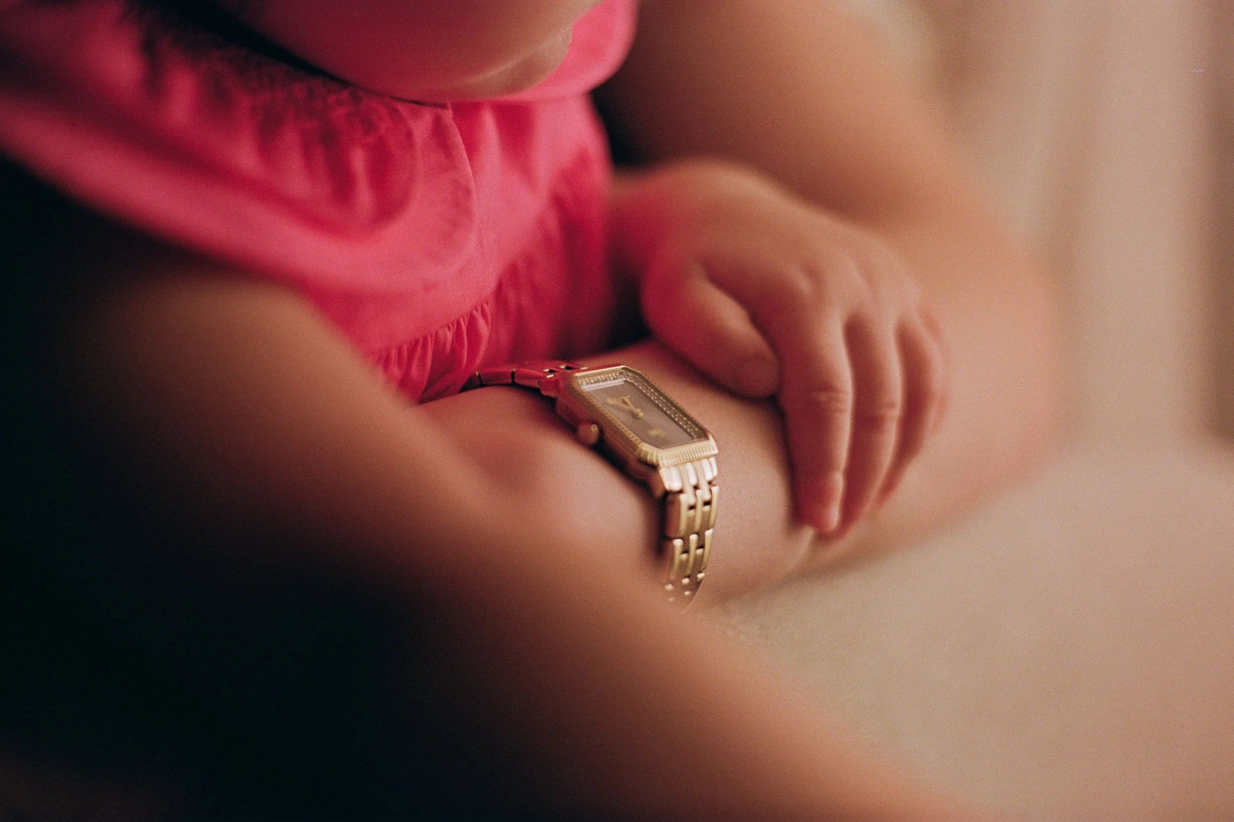 Close-up of a child's hand resting on their arm, wearing a gold watch, with a pink garment visible.