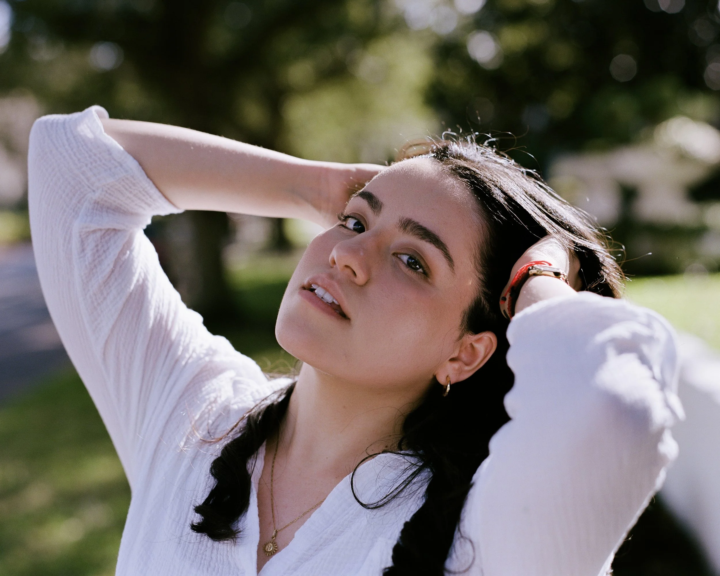 Close-up of a woman wearing a yellow floral dress, gold necklace with a square pendant, and sunglasses, with her hand near her head, outdoors during daytime.