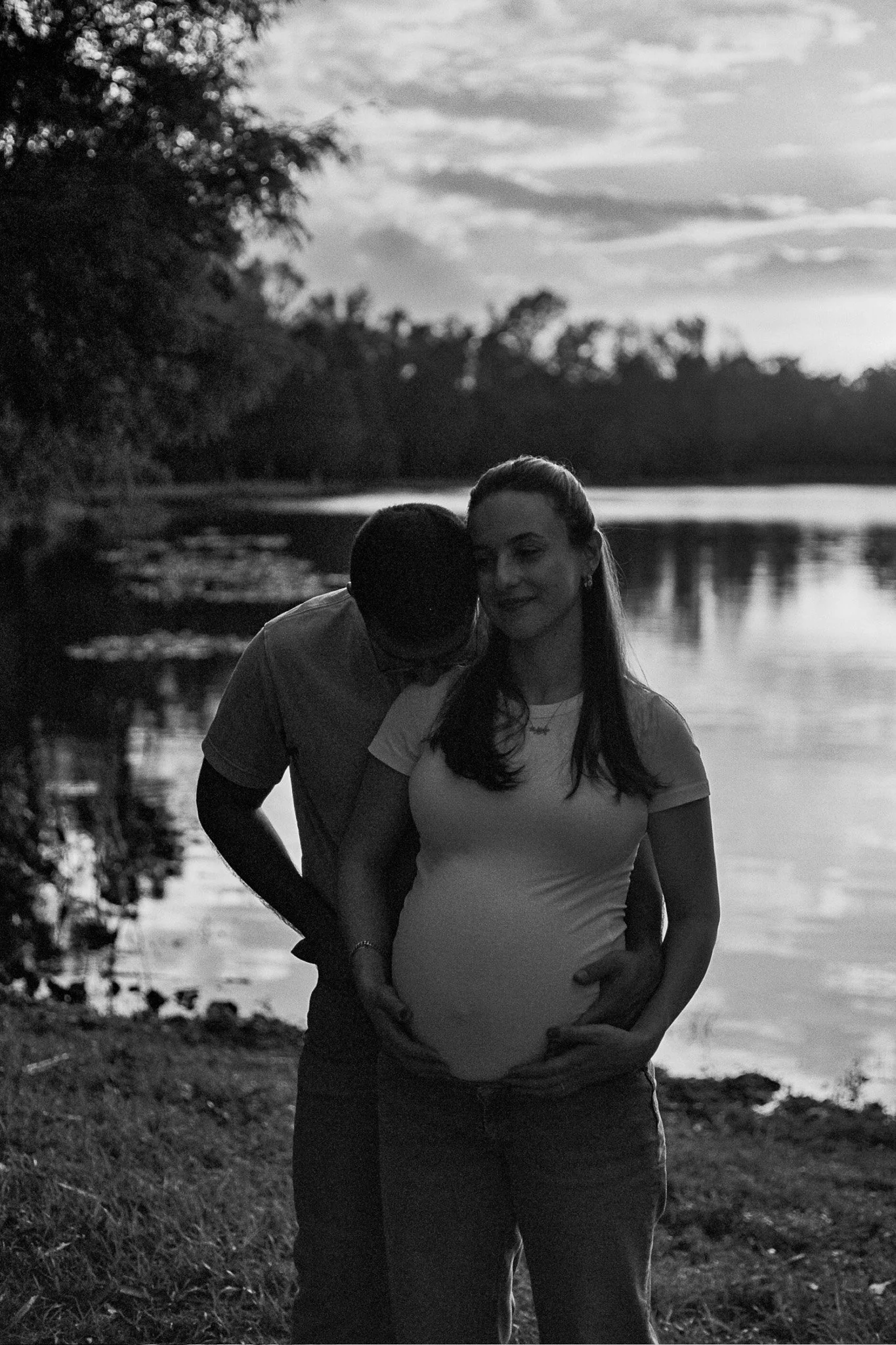 A pregnant woman standing by the water under a cloudy sky, with a man leaning in to kiss her head, during sunset.