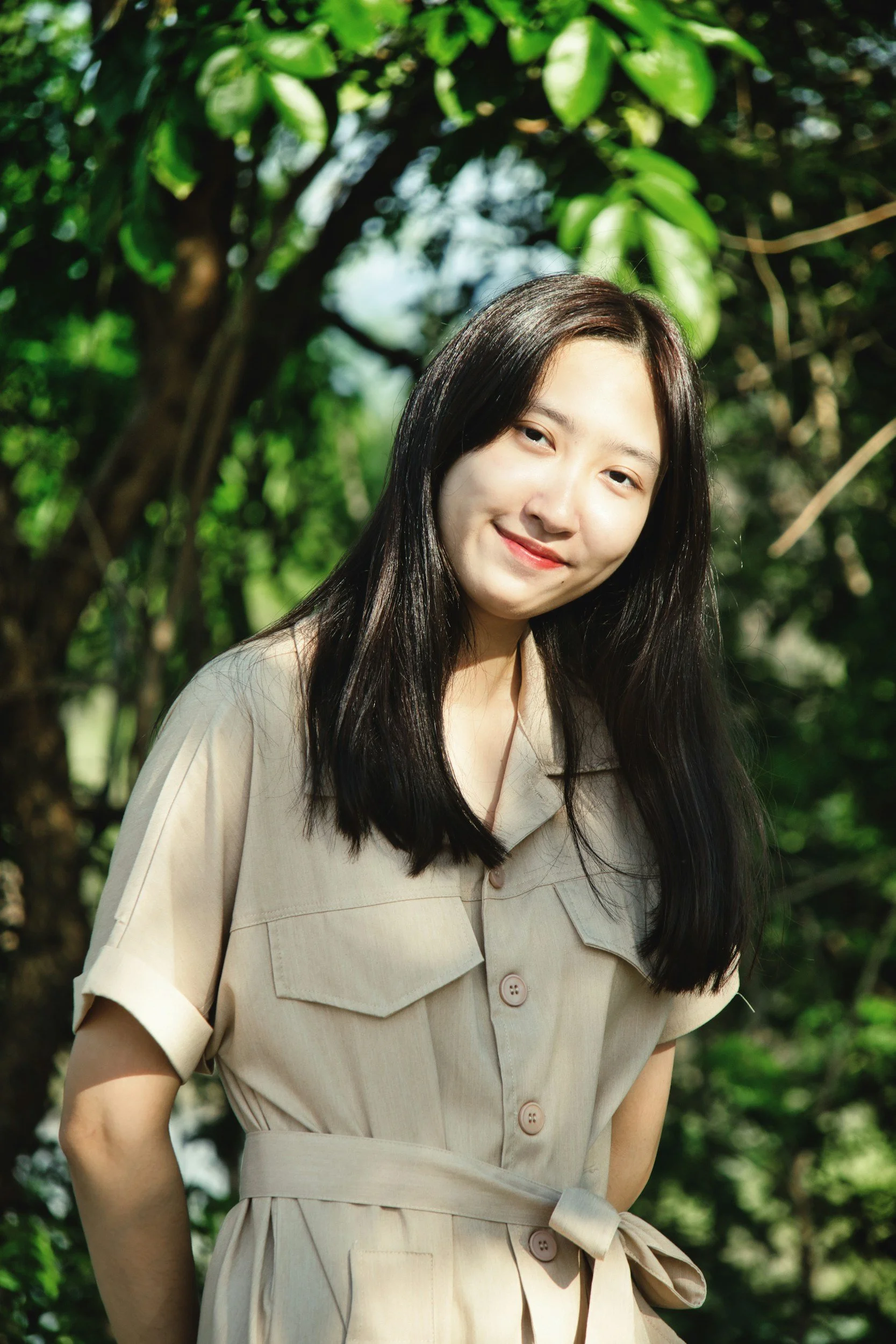 A young woman with long black hair smiling outdoors, wearing a beige dress with rolled-up sleeves and a waist belt, standing among green trees and foliage with sunlight filtering through.