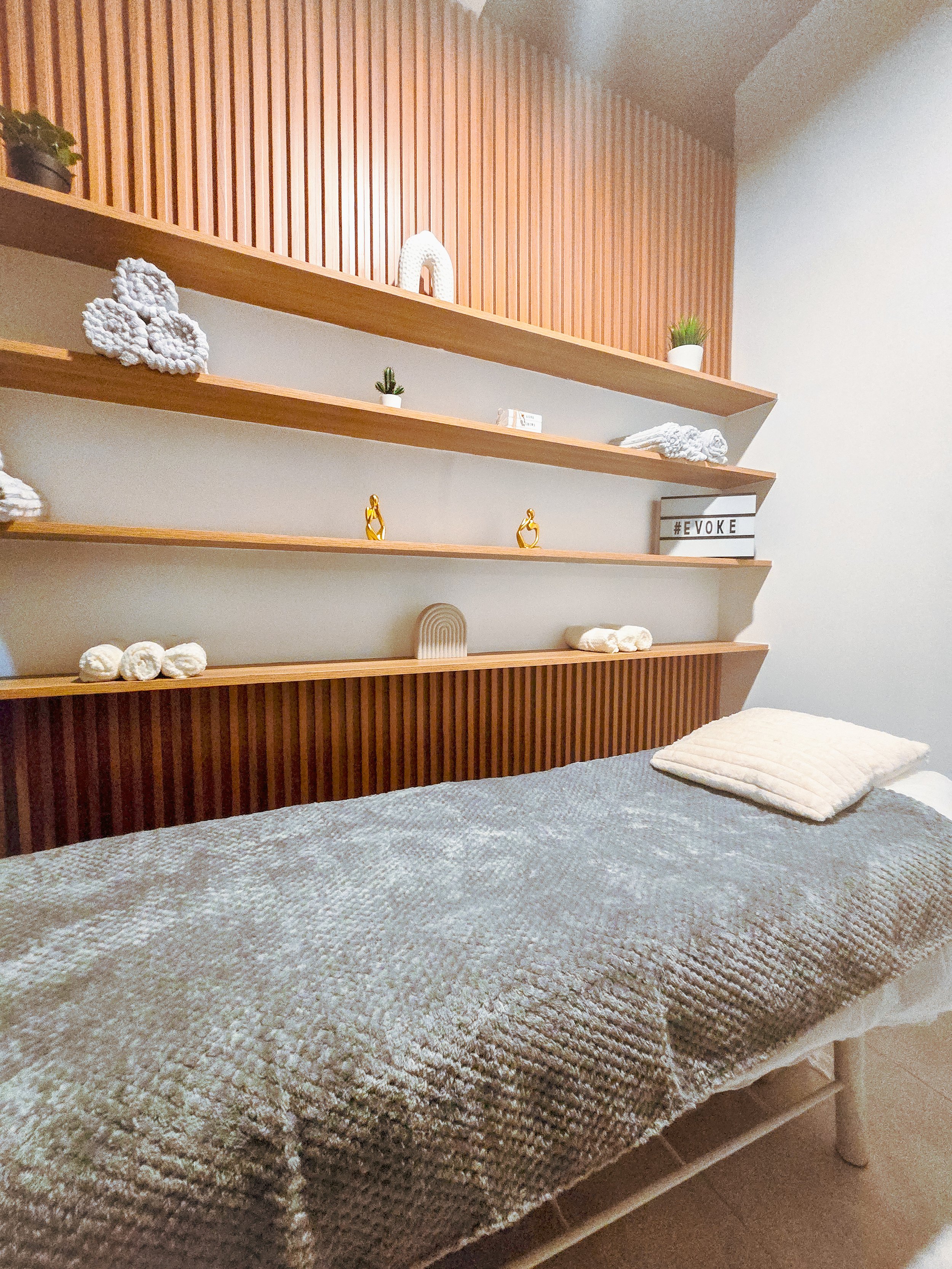 A massage therapy room with a bed covered in a textured gray blanket and a folded towel at the head, against a wooden-paneled wall with decorative shelves holding small plants, stones, and a lightbox with the word '#EVOKE'.