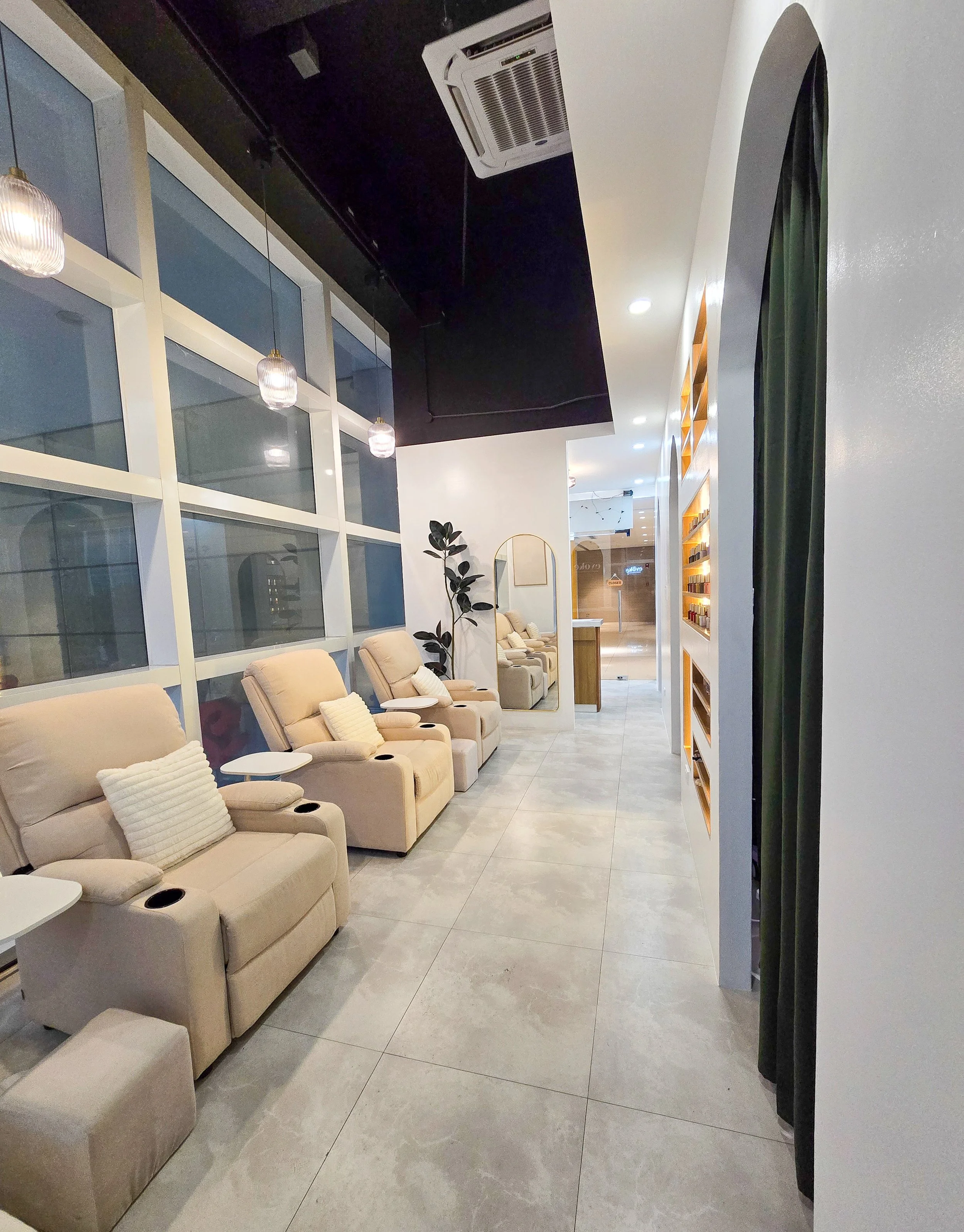 A waiting area with beige recliner chairs, small white side tables, a large mirror, potted plants, and a wall with glass windows and hanging pendant lights.