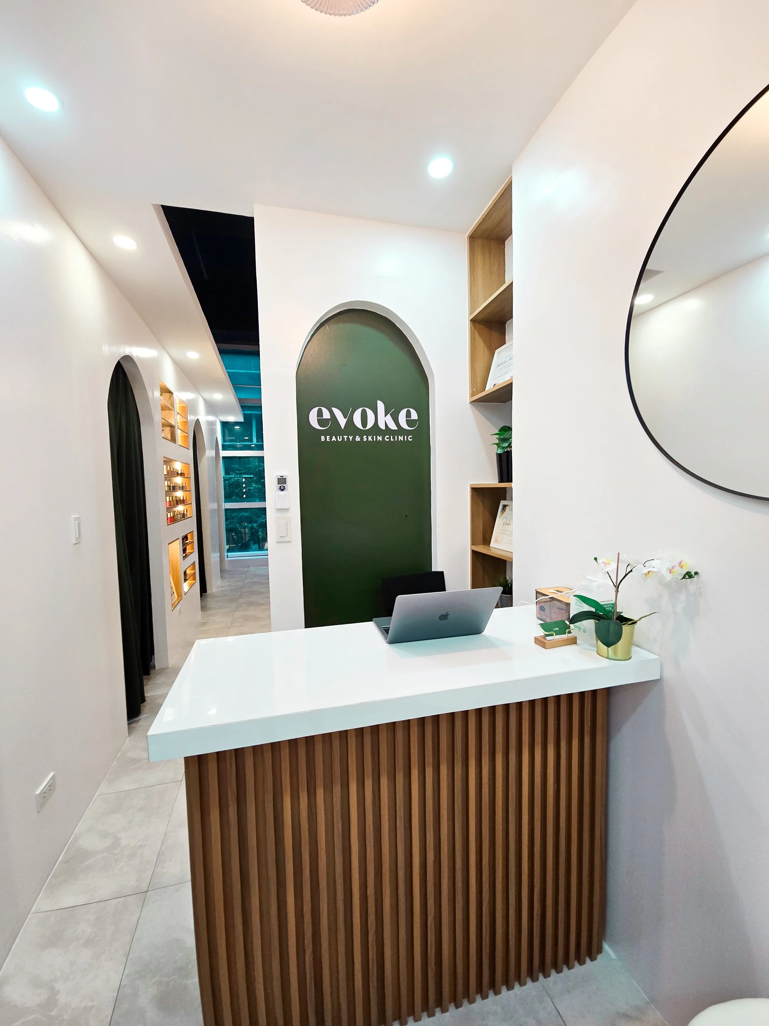 Reception area of a beauty and skin clinic with a white counter, a laptop, a mirror, and decorative plants, with the clinic's sign 'evoke' on a dark green wall.