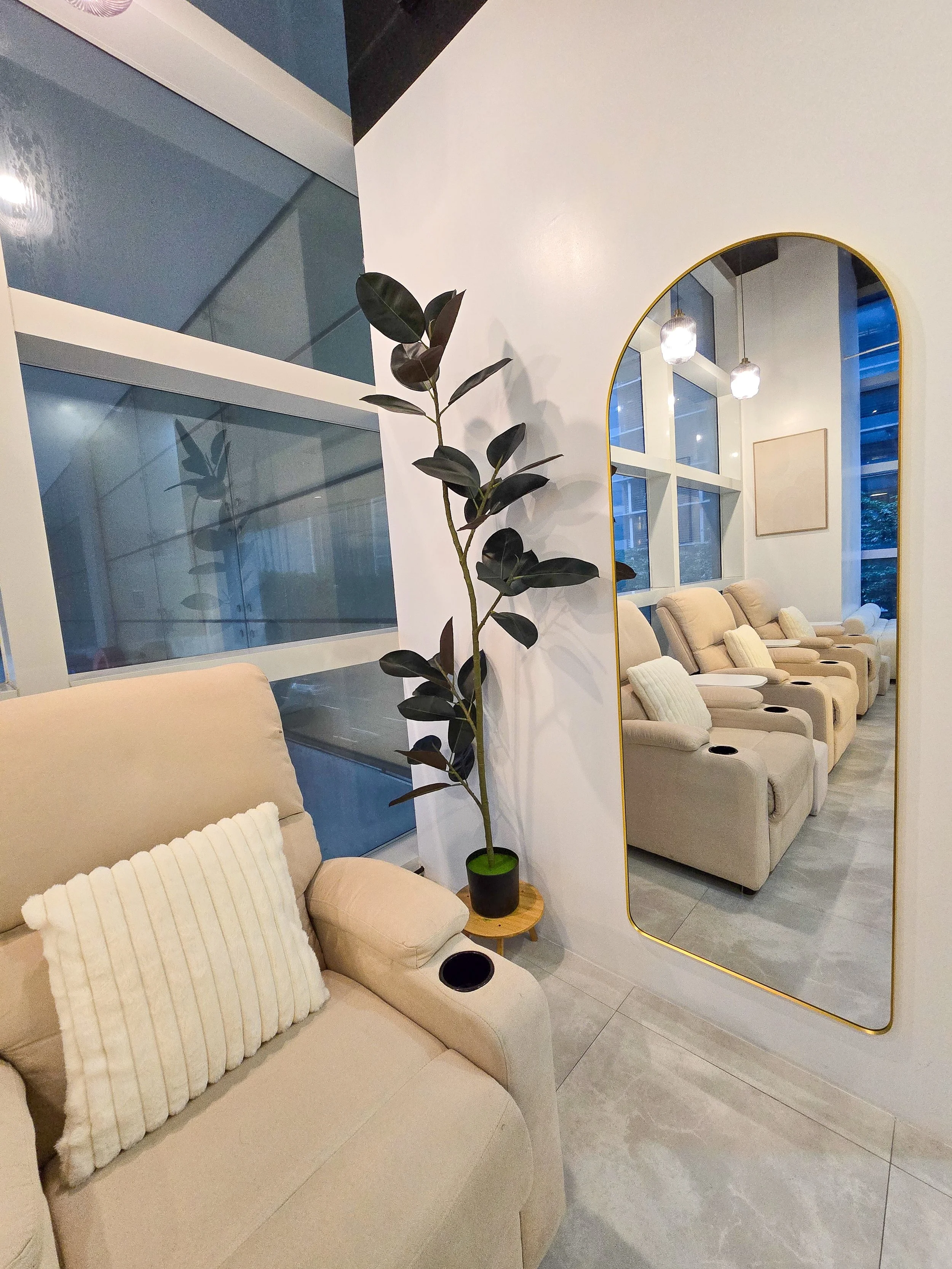 A cozy waiting area with beige recliner chairs, a large mirror with a gold frame, a tall potted plant, and a windowed wall in the background.