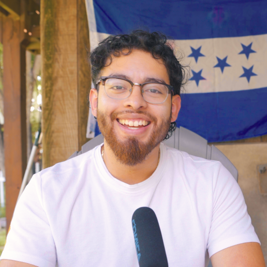 A smiling man with glasses and facial hair sitting outdoors, holding a microphone and wearing a white t-shirt, with a blue and white star-spangled flag hanging behind him.