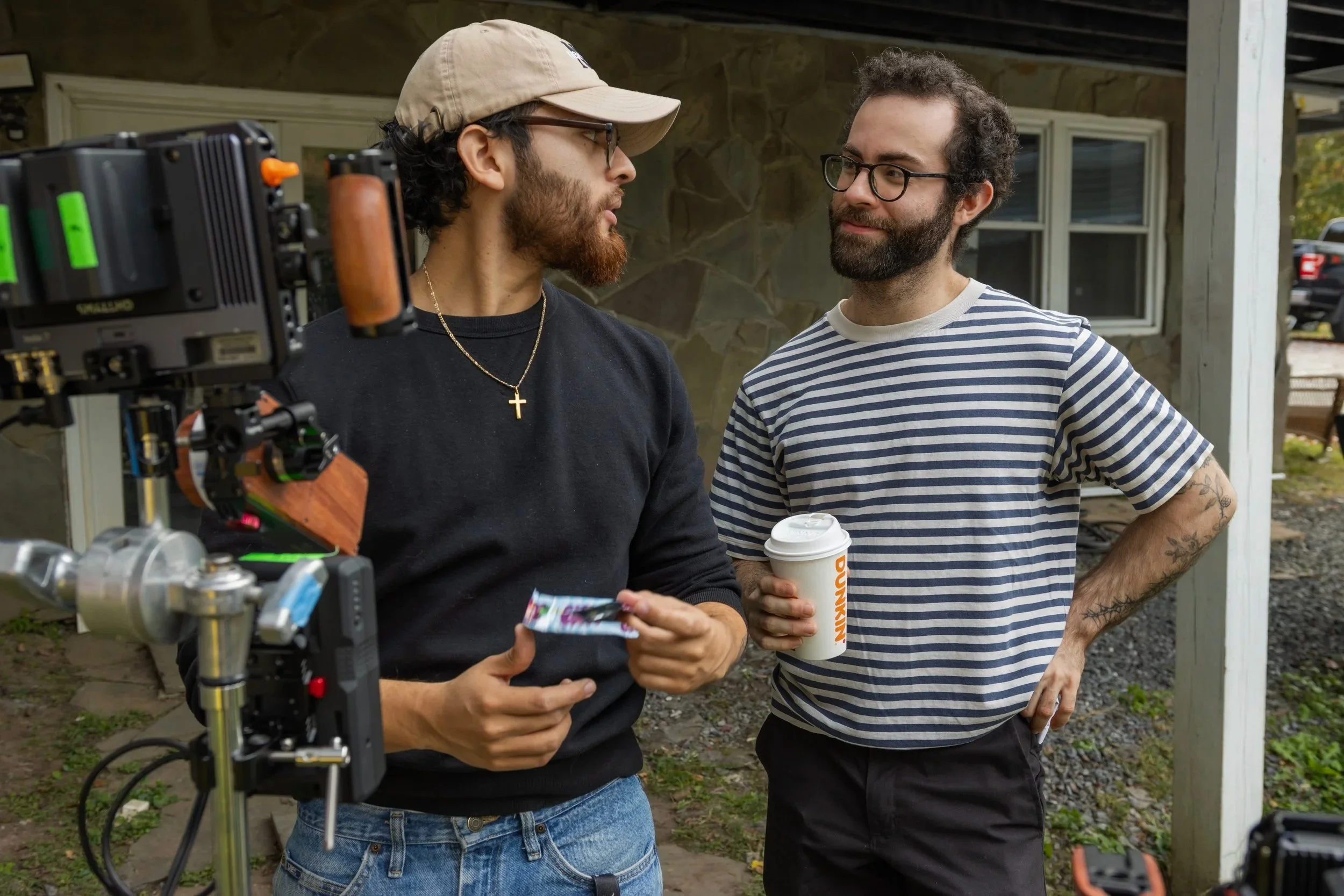 Two men having a conversation outdoors with film equipment in the foreground. One wearing a black sweatshirt, glasses, a beige cap, and a cross necklace, holding a colorful card. The other, in a striped shirt, glasses, and holding a Dunkin' coffee cup, has tattoos on his arm.