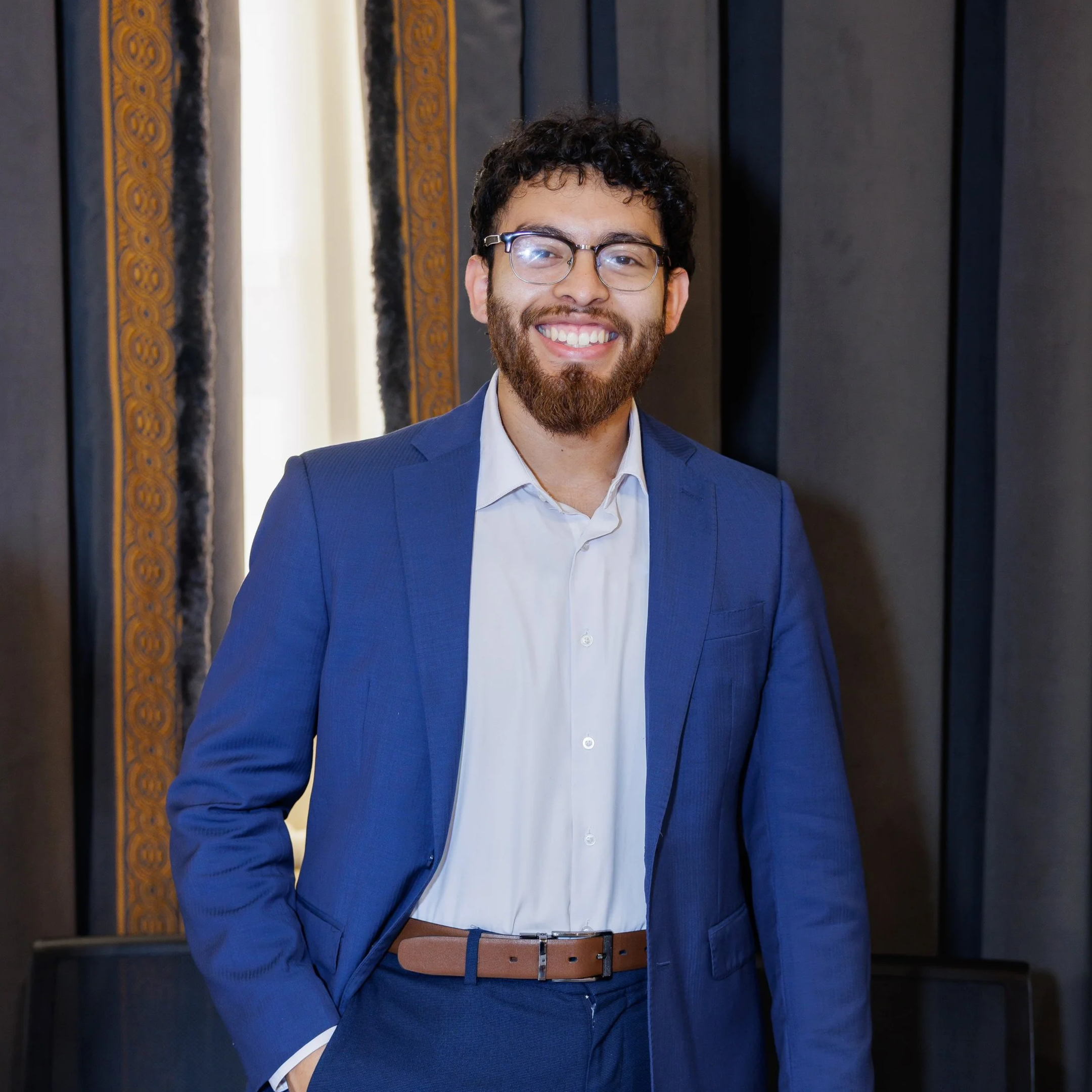 A man with curly dark hair, glasses, and a beard smiling wearing a blue suit jacket, white shirt, and a brown belt, standing against a dark background with decorative borders.
