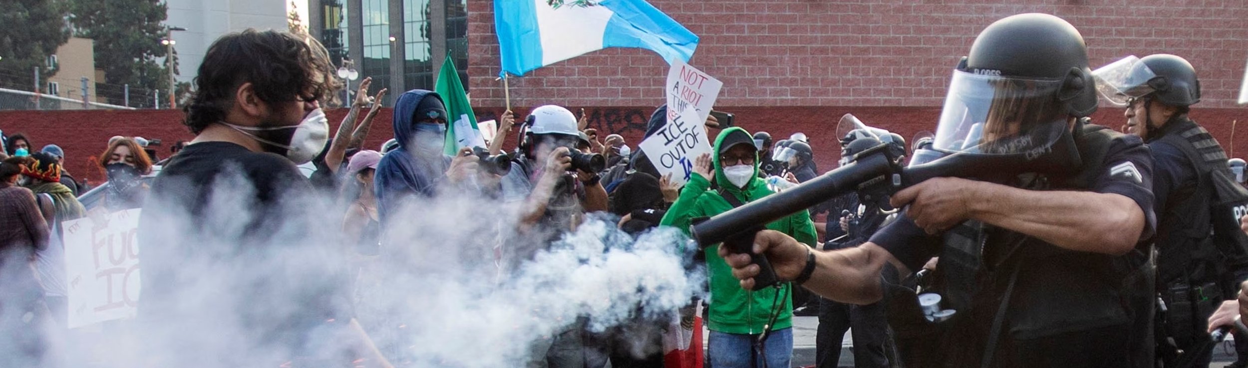 A protest or demonstration with a large crowd of people, some holding signs and flags, facing police officers in riot gear. One police officer is aiming a gun at the crowd, with smoke or tear gas in the foreground.