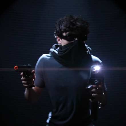 Person wearing a hoodie and mask, holding two guns, in a dark setting.