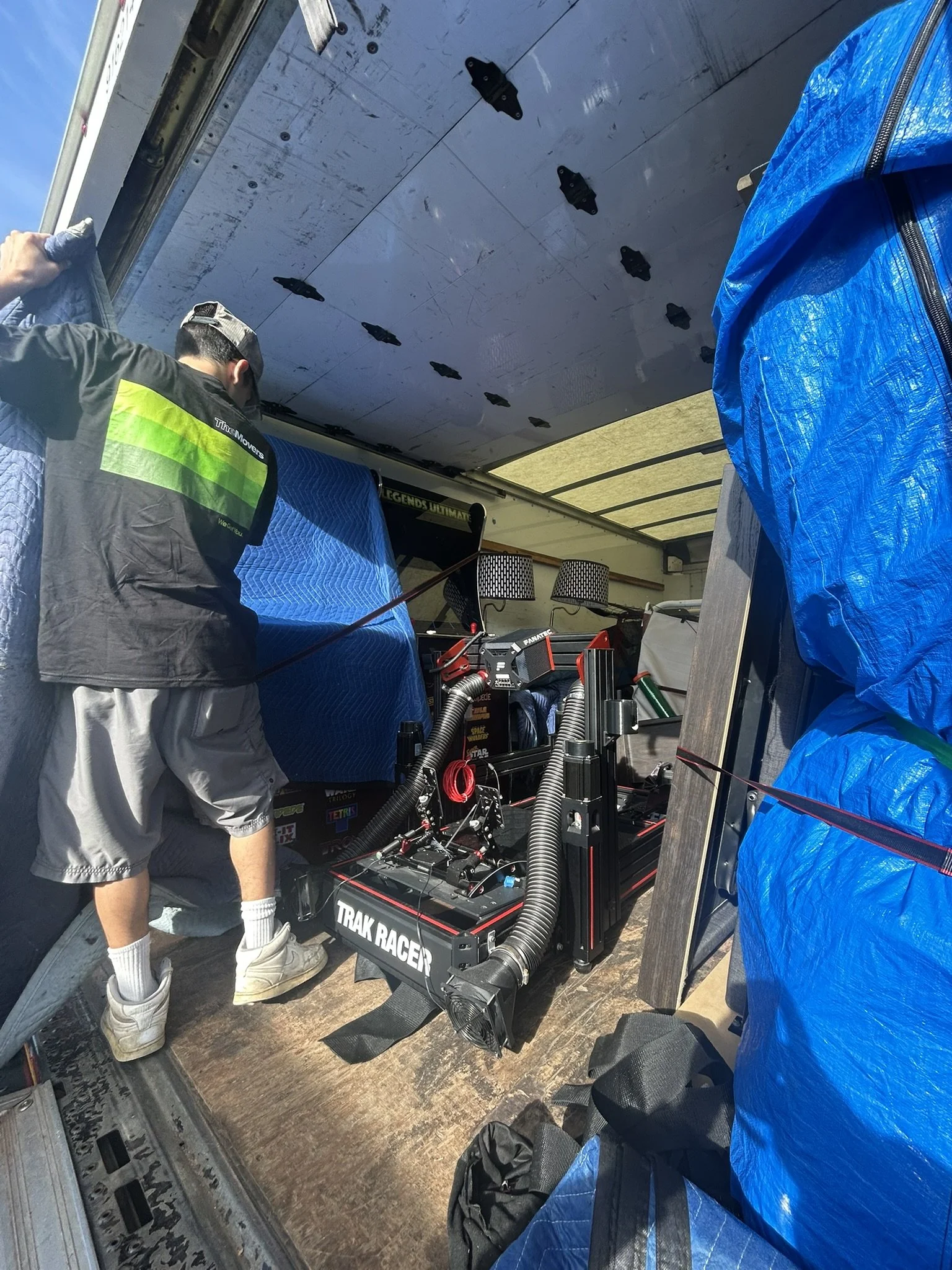 A person working inside a moving truck, surrounded by blue tarps and various equipment, including a racing simulator named 'TRAK RAGER'. Pearl City.