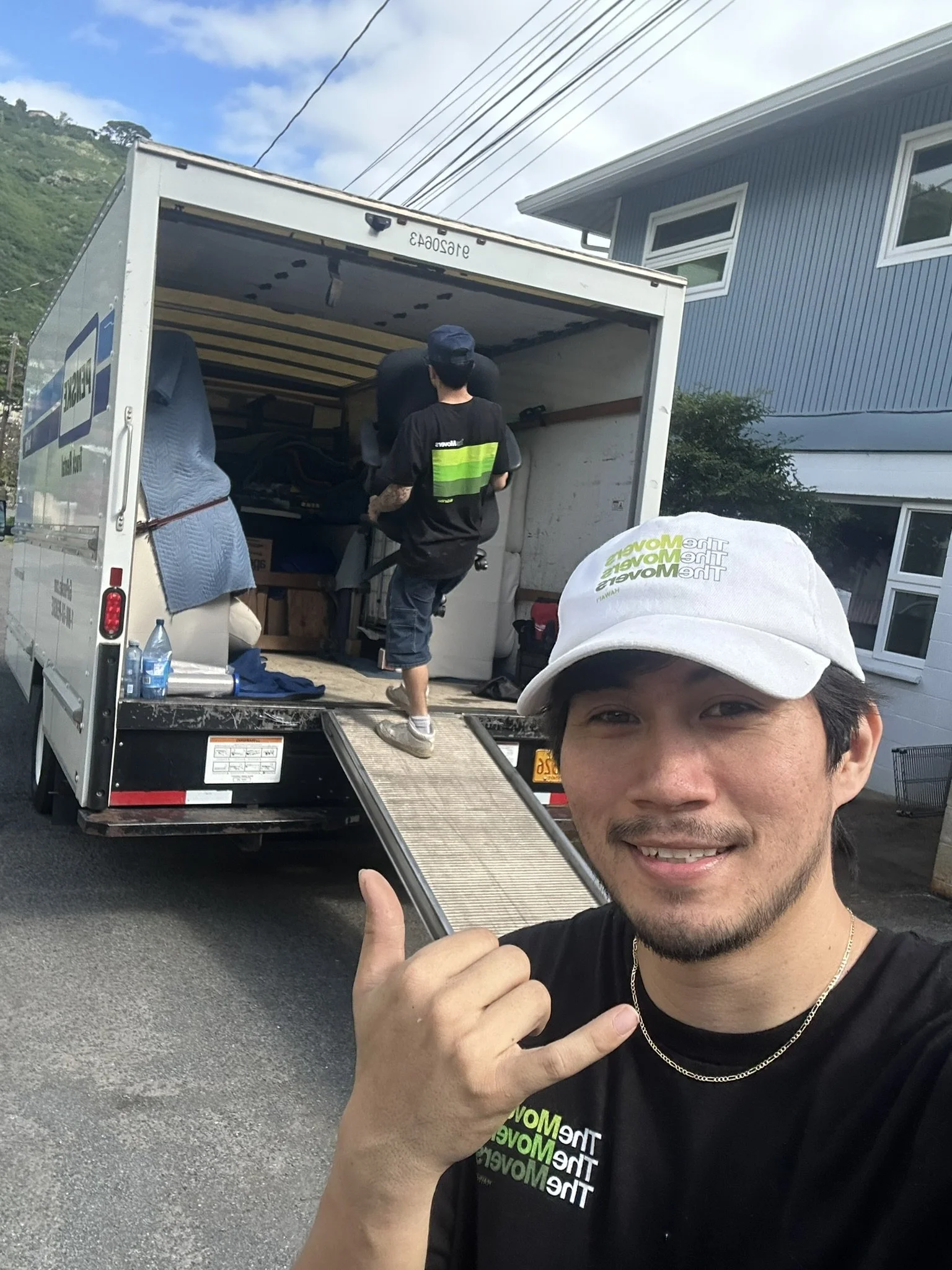 A young man smiling and making a hang loose, shaka, gesture in front of an open moving truck, with another person loading items inside. The scene takes place outdoors in a residential area with a blue house and a hill in the background. Manoa.
