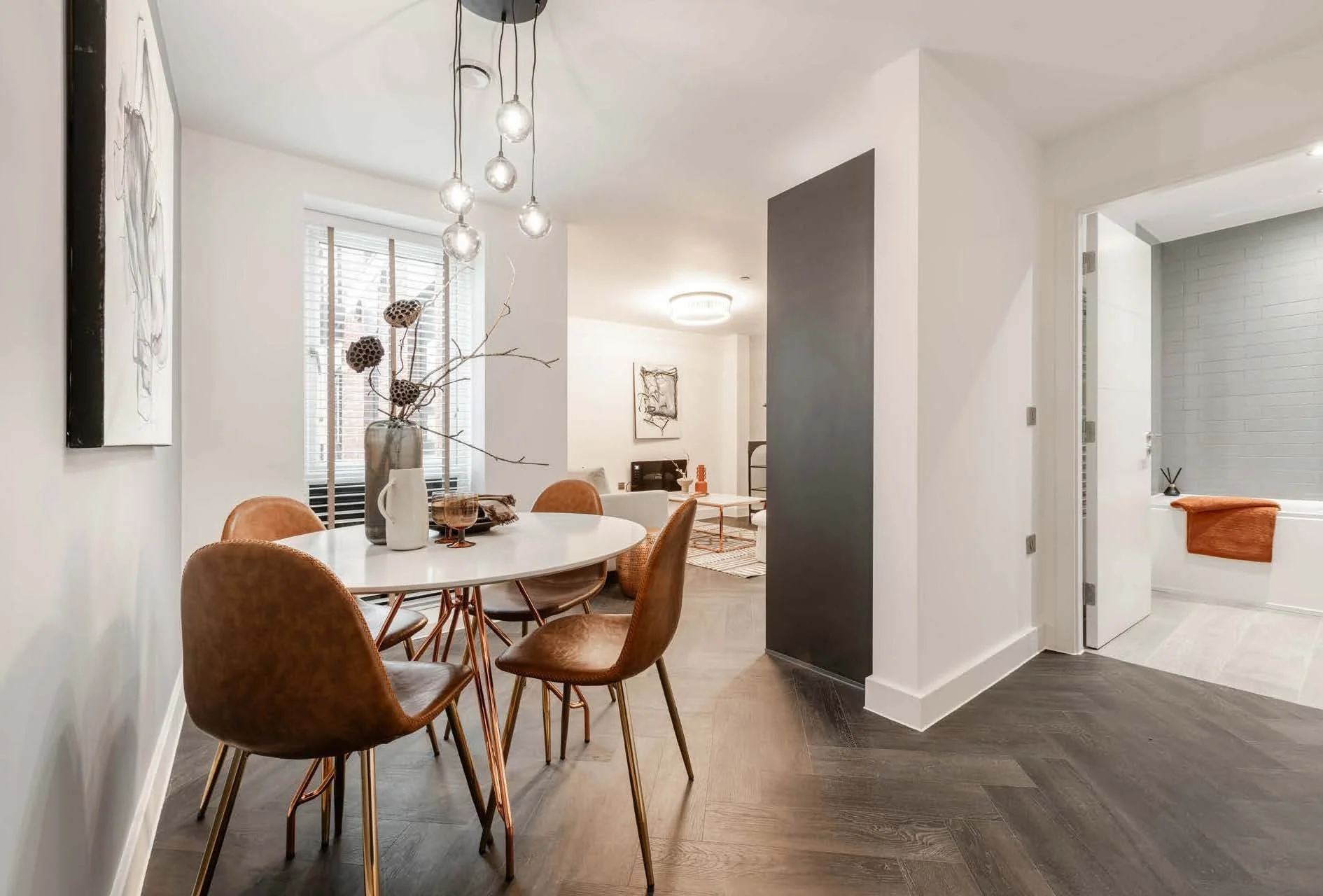 Dining area with a white oval table, four brown leather chairs, a vase with dried flowers, and a window with blinds. The background shows a living room and a bathroom with a bathtub and orange towel.