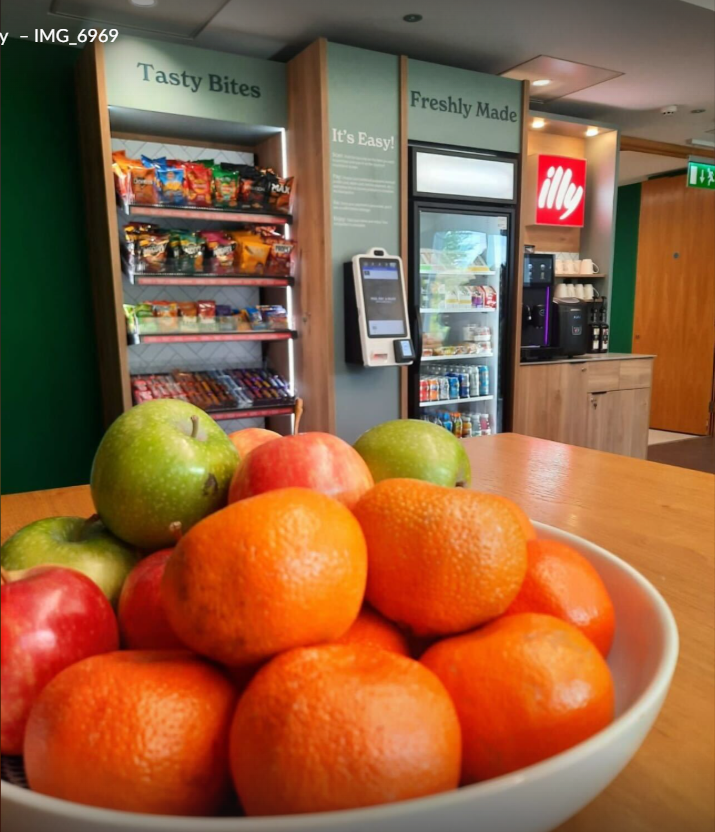 Een witte kom met sinaasappels en appels op een houten tafel, met een achtergrond van snacks, een zelfbedieningskast, een koffiezetapparaat en een zakje illy koffie in een koffieautomaat in een horecagelegenheid.