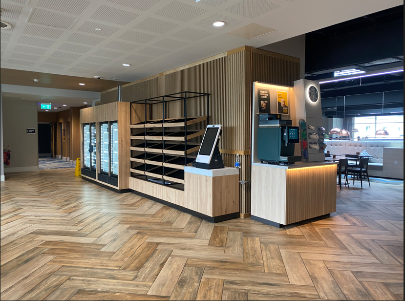 Verdieping met een zelfbedieningskast en gekoelde rekken in een hotel of restaurant, met een touchscreen kiosk en een koffieautomaat, en zitplaatsen in de achtergrond.