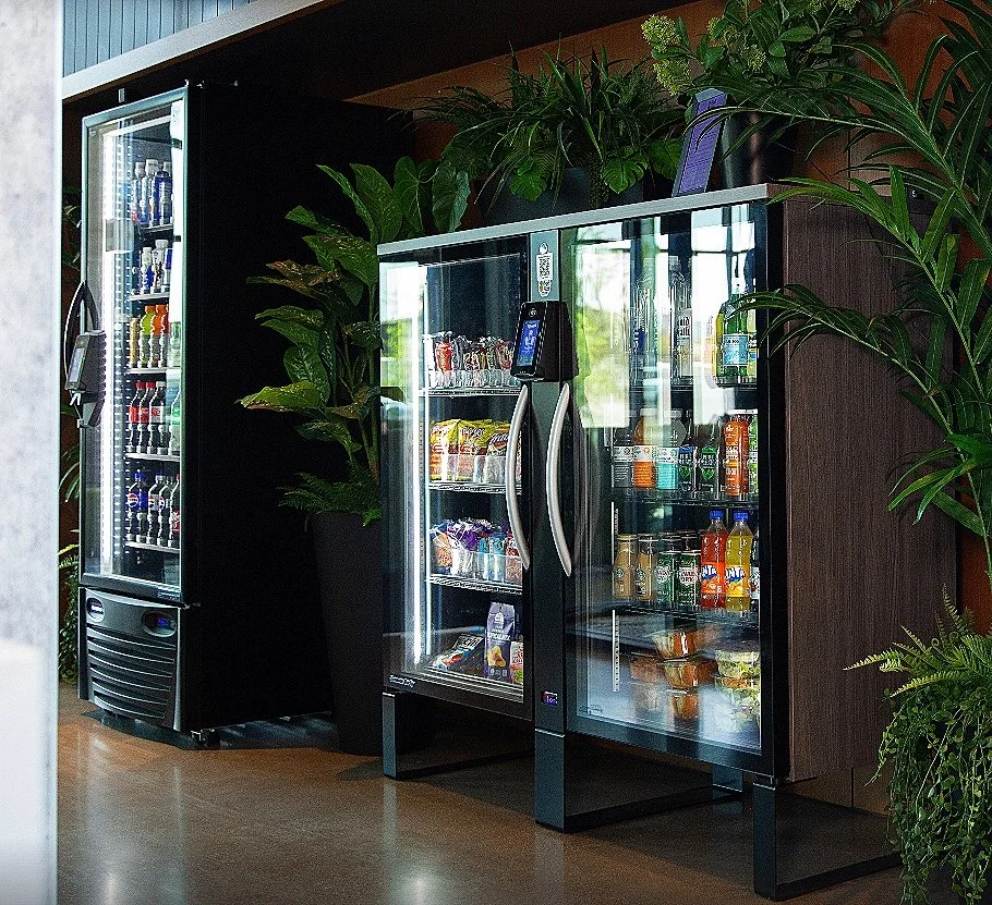 Vijf koelkasten gevuld met drankjes en snacks, omgeven door groene planten in een interieur met houten accenten.