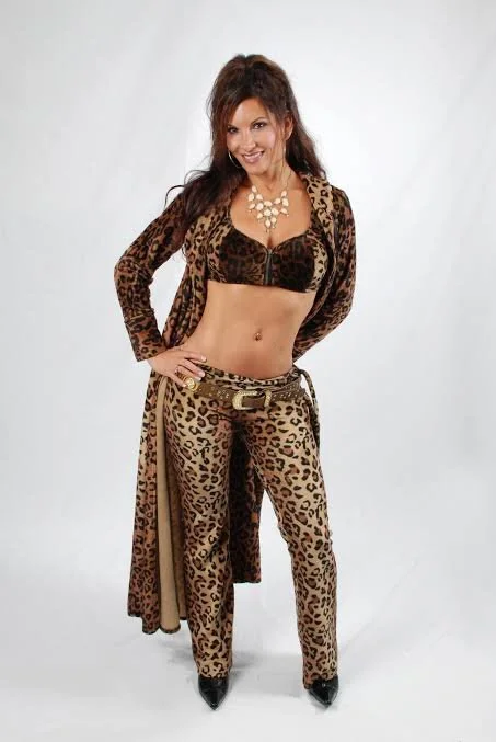 Woman standing in leopard print matching outfit, including pants, top, and long coat, with a necklace and high heels, posing with hands on hips against a plain white background.