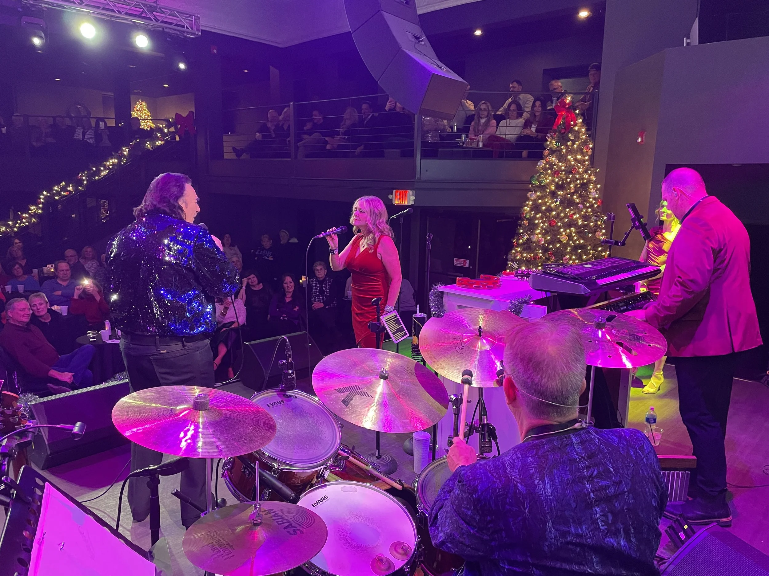 A live jazz performance during a holiday celebration, with a jazz band and singers on stage, decorated with Christmas lights and a Christmas tree, in a venue with audience sitting and standing, including a balcony with additional spectators.