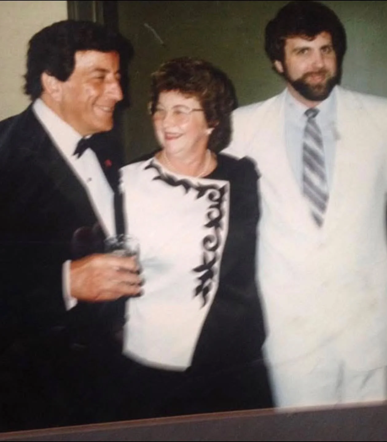 Three people at a social event: a man in a tuxedo holding a drink, a woman in a black-and-white dress, and a man in a white suit with a striped tie, smiling and talking.