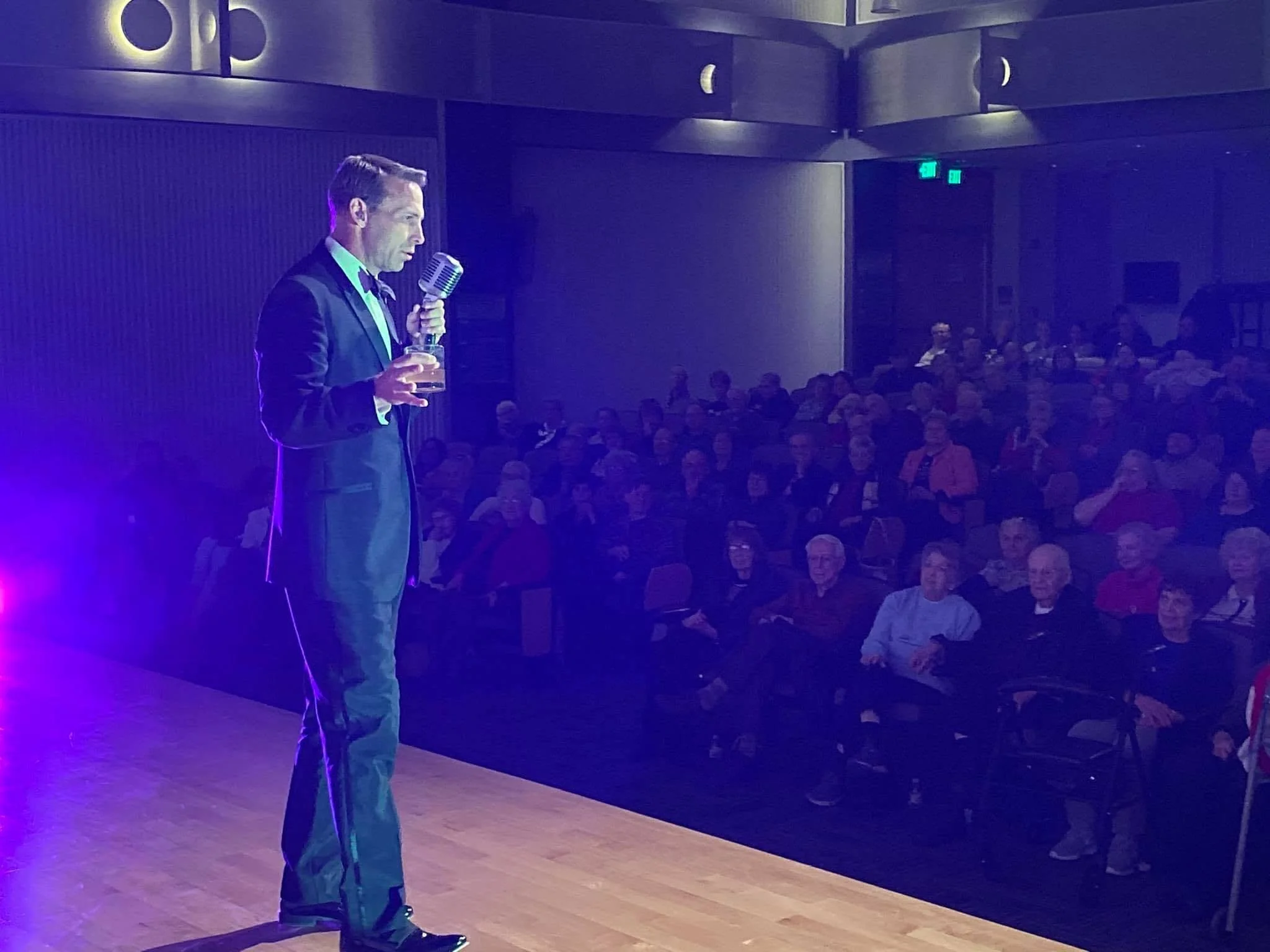 A man in a suit holding a microphone and a glass of drink, standing on stage in front of an audience in a dimly-lit room, with purple and blue lighting.