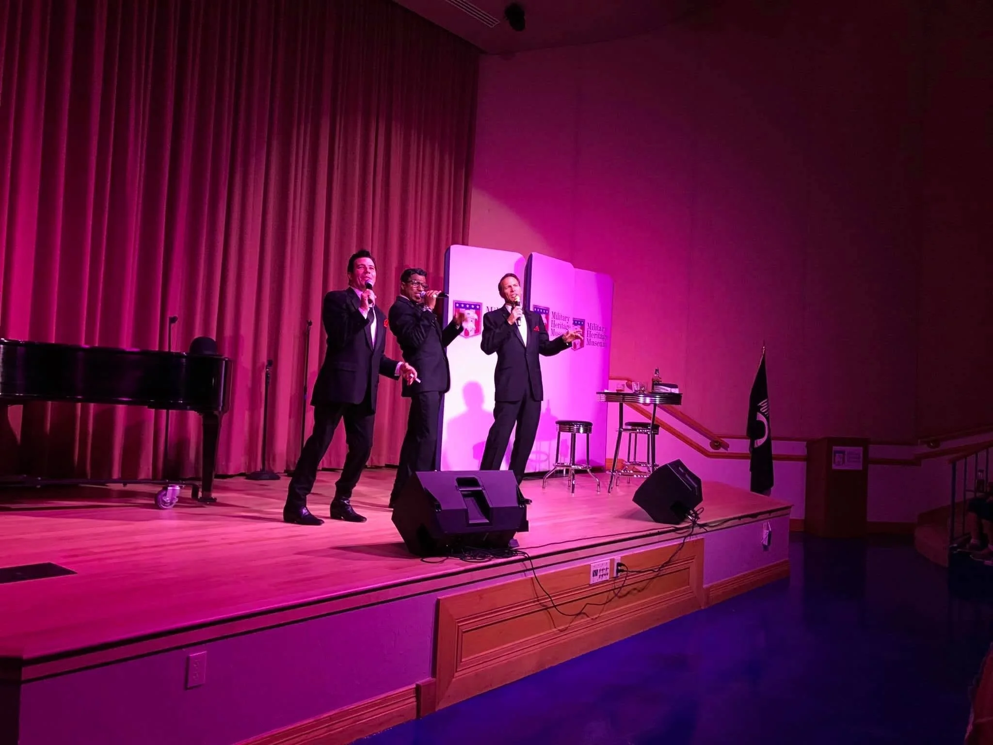 Three men in suits singing on a stage with a pink and purple backdrop, a black grand piano, and chairs.