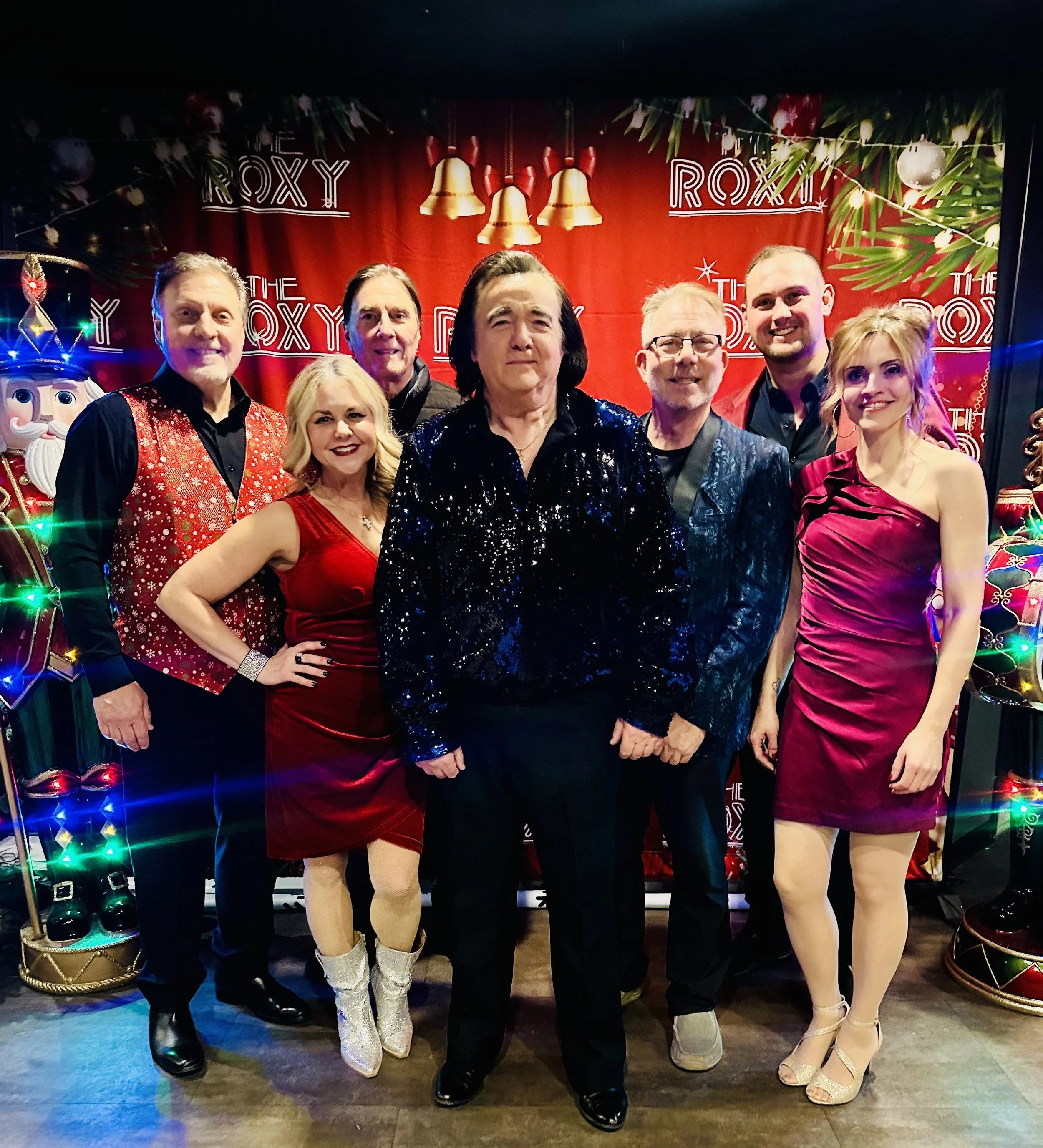 Group of seven people dressed in festive attire standing in front of a red background with holiday decorations and banner reading 'The Roxy'.