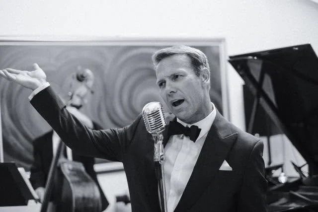 A man in a tuxedo singing or speaking into a vintage microphone, with a pianist visible in the background.