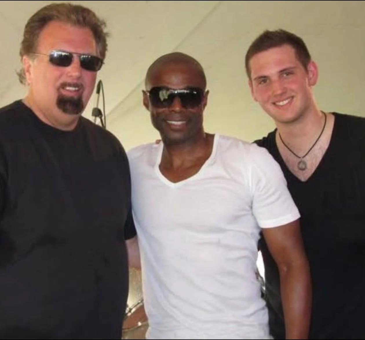 Three men smiling and standing close together indoors, wearing casual black and white t-shirts, with two of them wearing sunglasses.