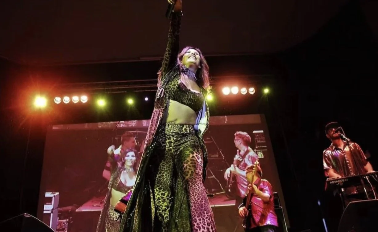 Performer on stage wearing a leopard print outfit with a long coat, smiling, with stage lights and a large screen in the background showing her image, a musician with a guitar, and a DJ with a turntable.