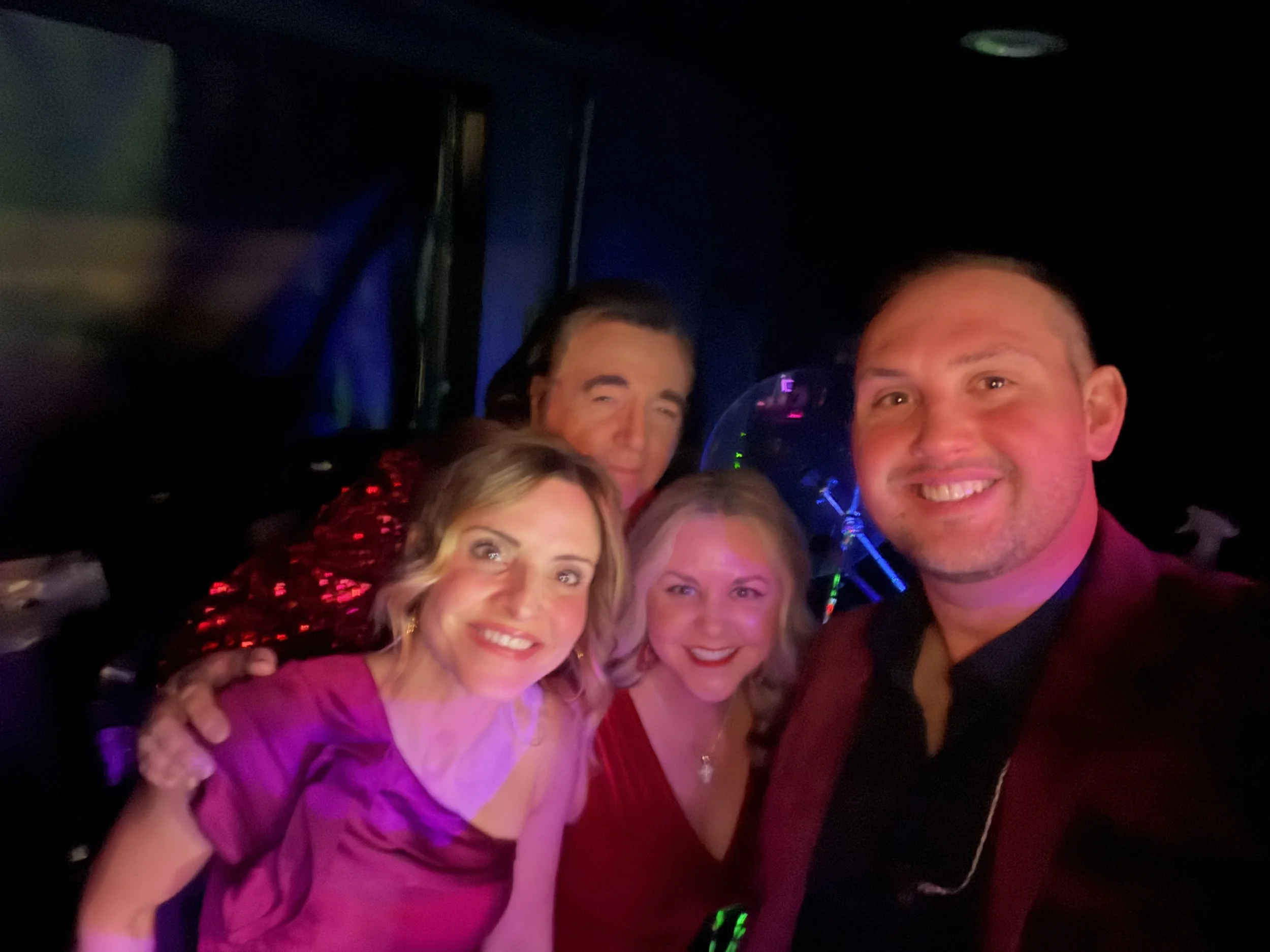 Group of five smiling people, three women and two men, close together, with a dark background and colorful lighting.